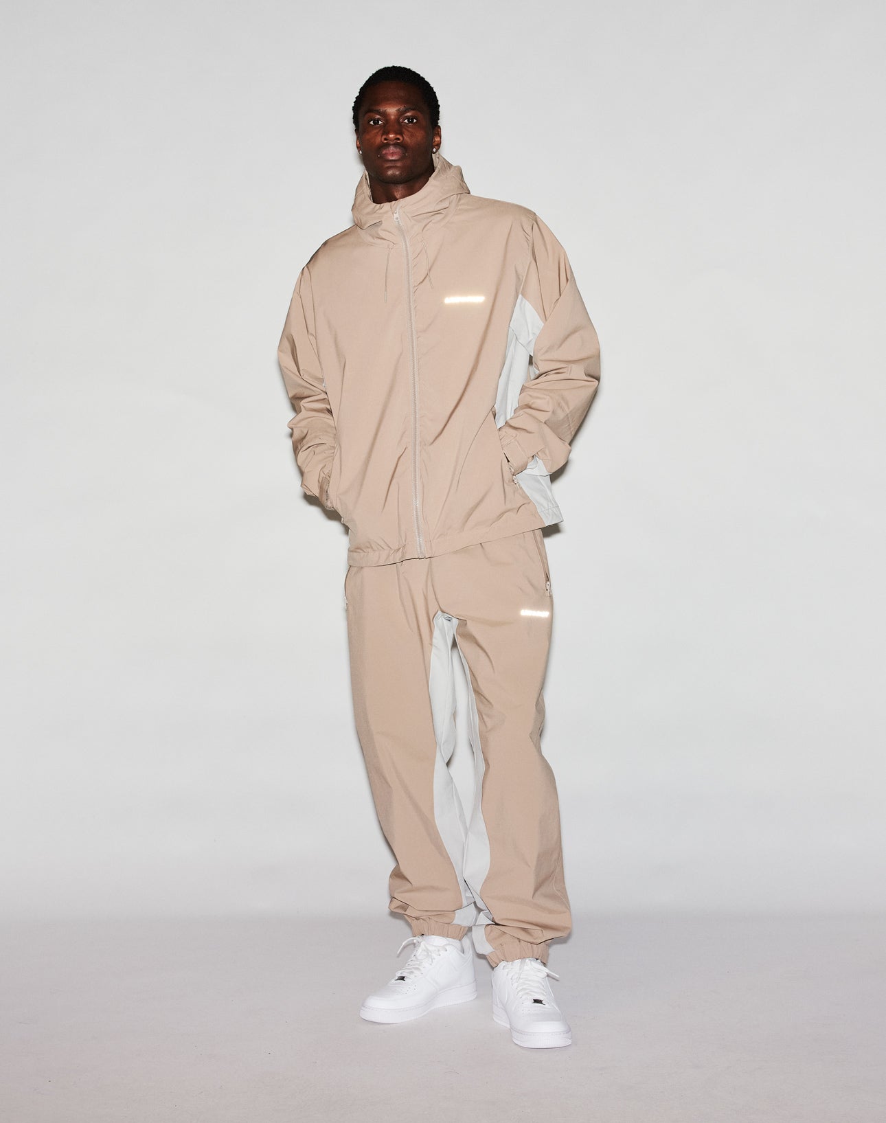 A man stands against a plain white background wearing the Basic Shell Jacket by LIVE FAST DIE YOUNG, an oversized beige and light gray hooded tracksuit with a wasserabweisender Reißverschluss, white sneakers, and his hands in his pockets.