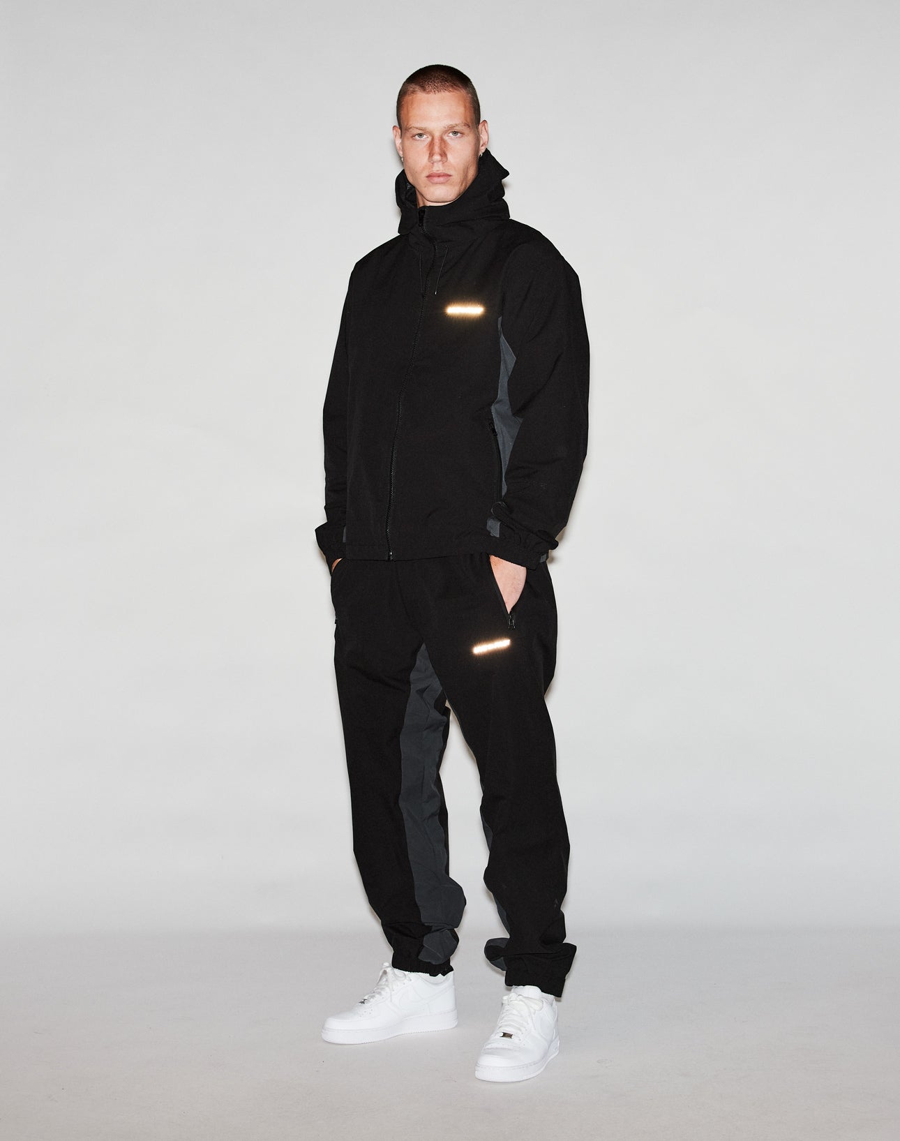 A person faces forward in a LIVE FAST DIE YOUNG Basic Shell Jacket with reflektierender Druck, black pants with reflective patches, and white sneakers against a plain light gray background.