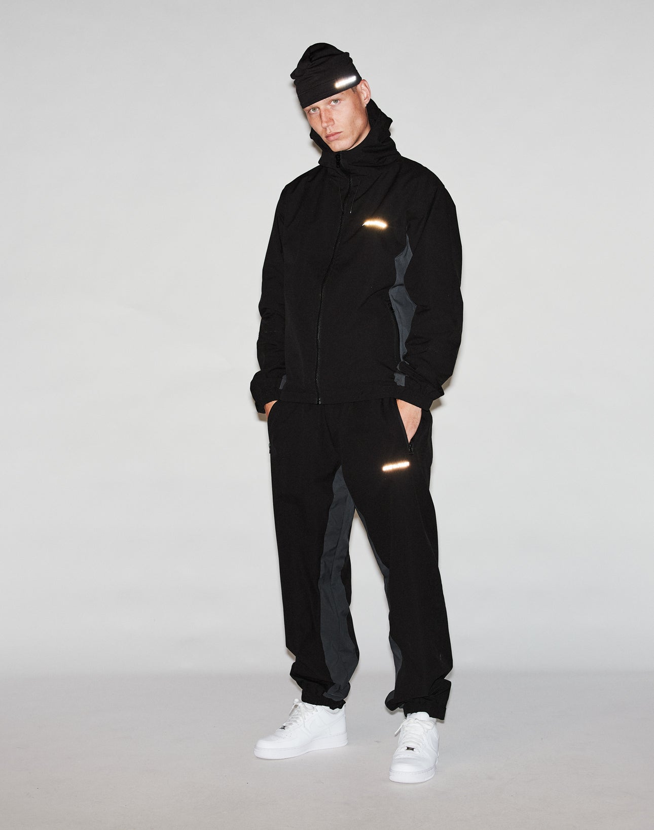 A person wearing a black oversize fit jacket, LIVE FAST DIE YOUNG Basic Shell Pants with reflektierender Druck and elastikbund mit Kordelzug, a black beanie, and white sneakers stands against a light gray background.