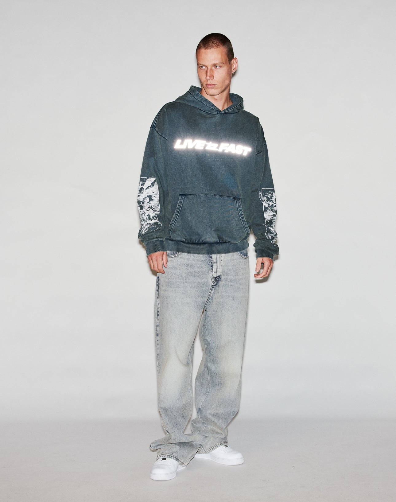 Someone in a dark LIVE FAST DIE YOUNG LFDY Window Hooded with LIVESTRAT front print and patterned sleeves poses against a plain background, styled with baggy light-wash jeans and white sneakers for an oversized look.
