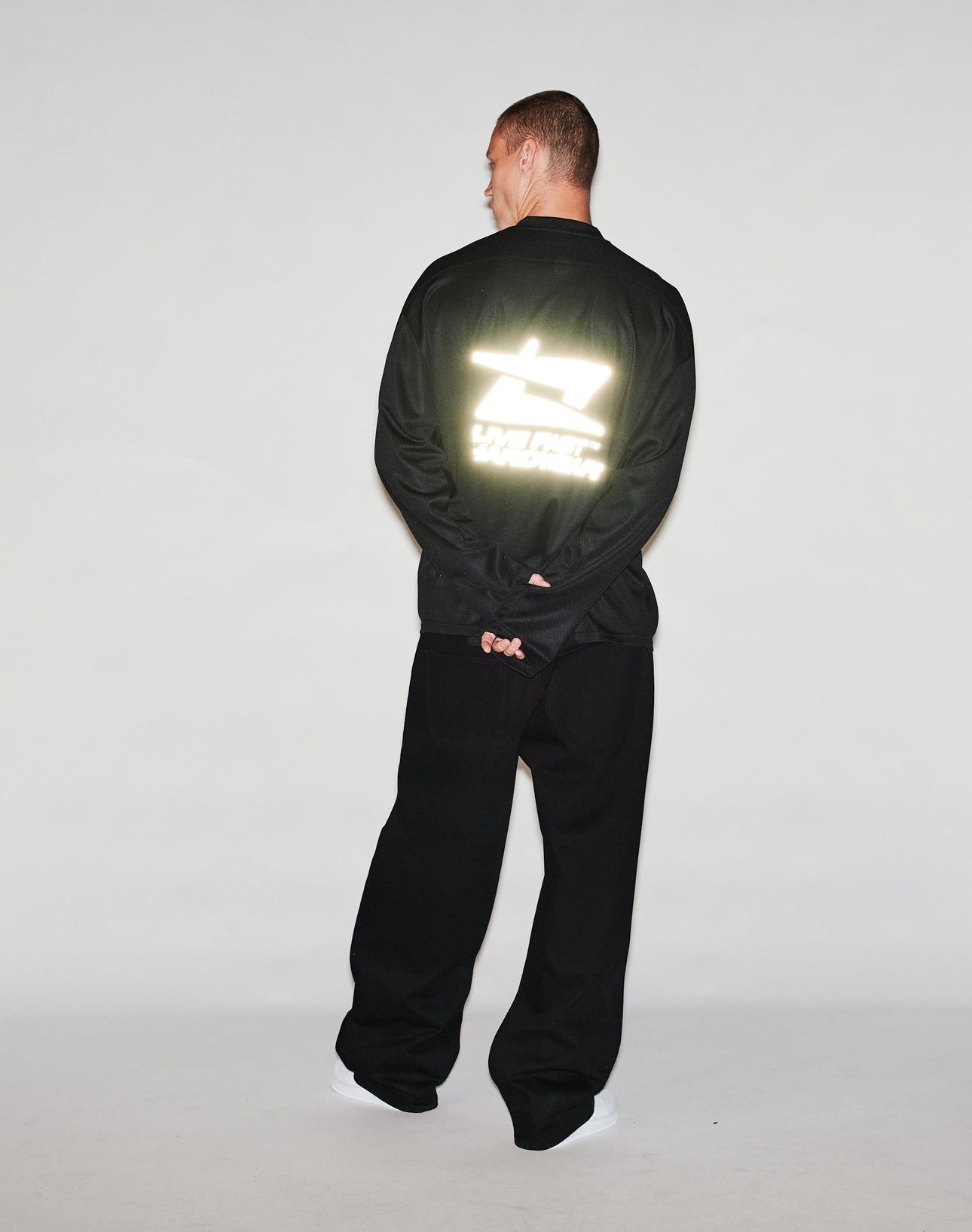 A person stands facing away with hands behind their back, wearing the LIVE FAST DIE YOUNG Live Fast Jersey in black. A bright, reflective print and text appear on the back of the top against a plain light background.