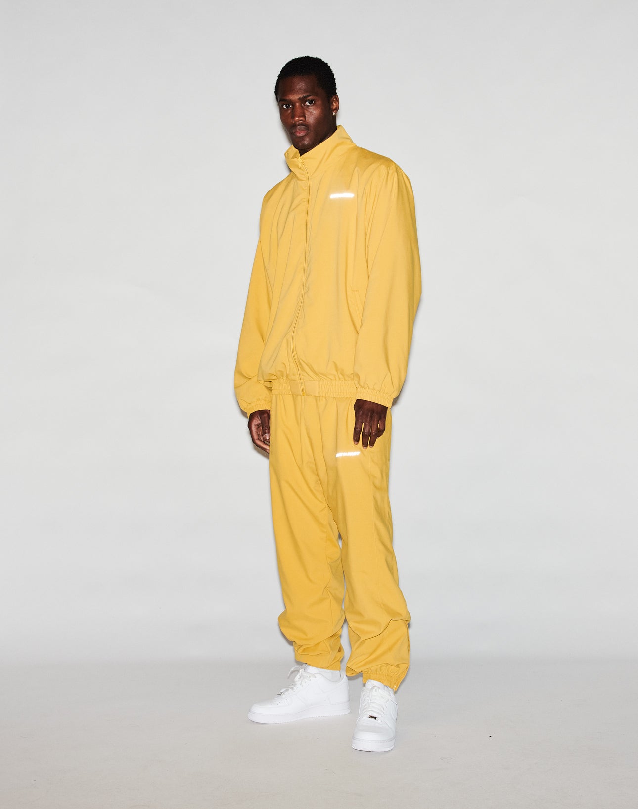 A man faces the camera against a plain gray background, wearing LIVE FAST DIE YOUNGs LFDY Trackpants—a loose-fit yellow polyester tracksuit with high collar, elastic waistband, white sneakers, and reflective print accents.