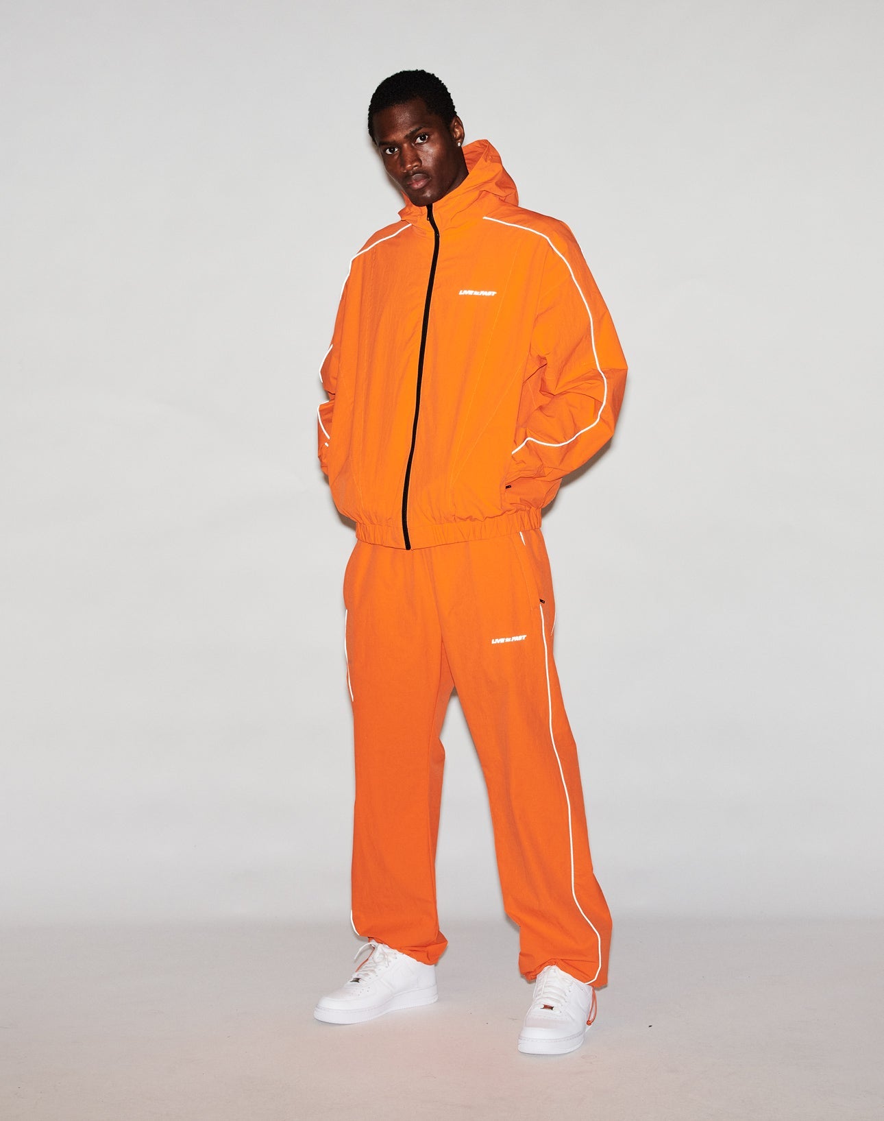 A person wears the LIVE FAST DIE YOUNG Live Fast Trackjacket, featuring bright orange color, reflektierender Druck, and elastische Bündchen, paired with white sneakers. They stand against a plain background with hands in the jacket pockets.