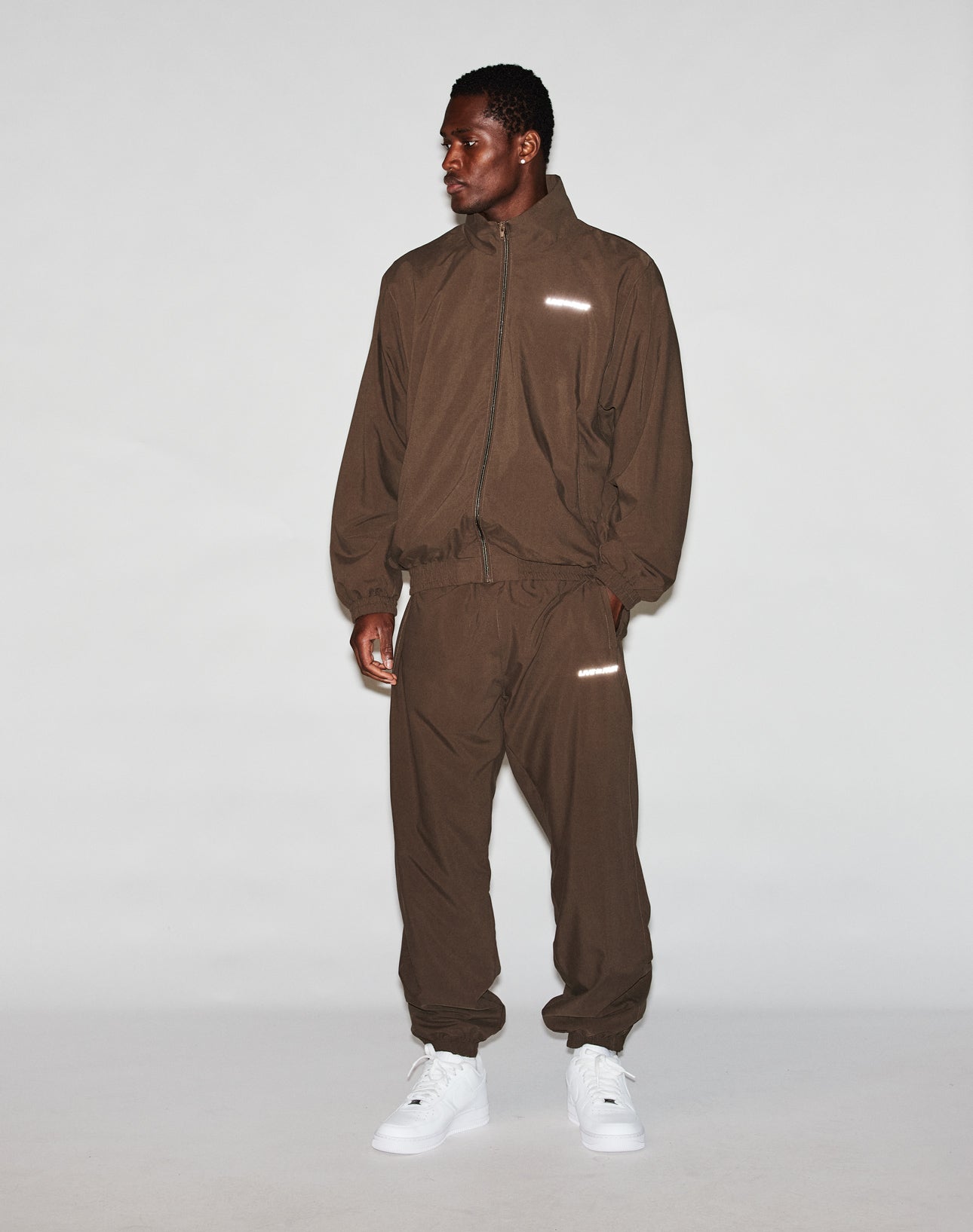 A man stands against a plain background wearing LIVE FAST DIE YOUNG LFDY Trackpants with a loose brown polyester zip-up jacket and elastic cuffs, paired with white sneakers. He looks to the side with his hands relaxed by his sides.