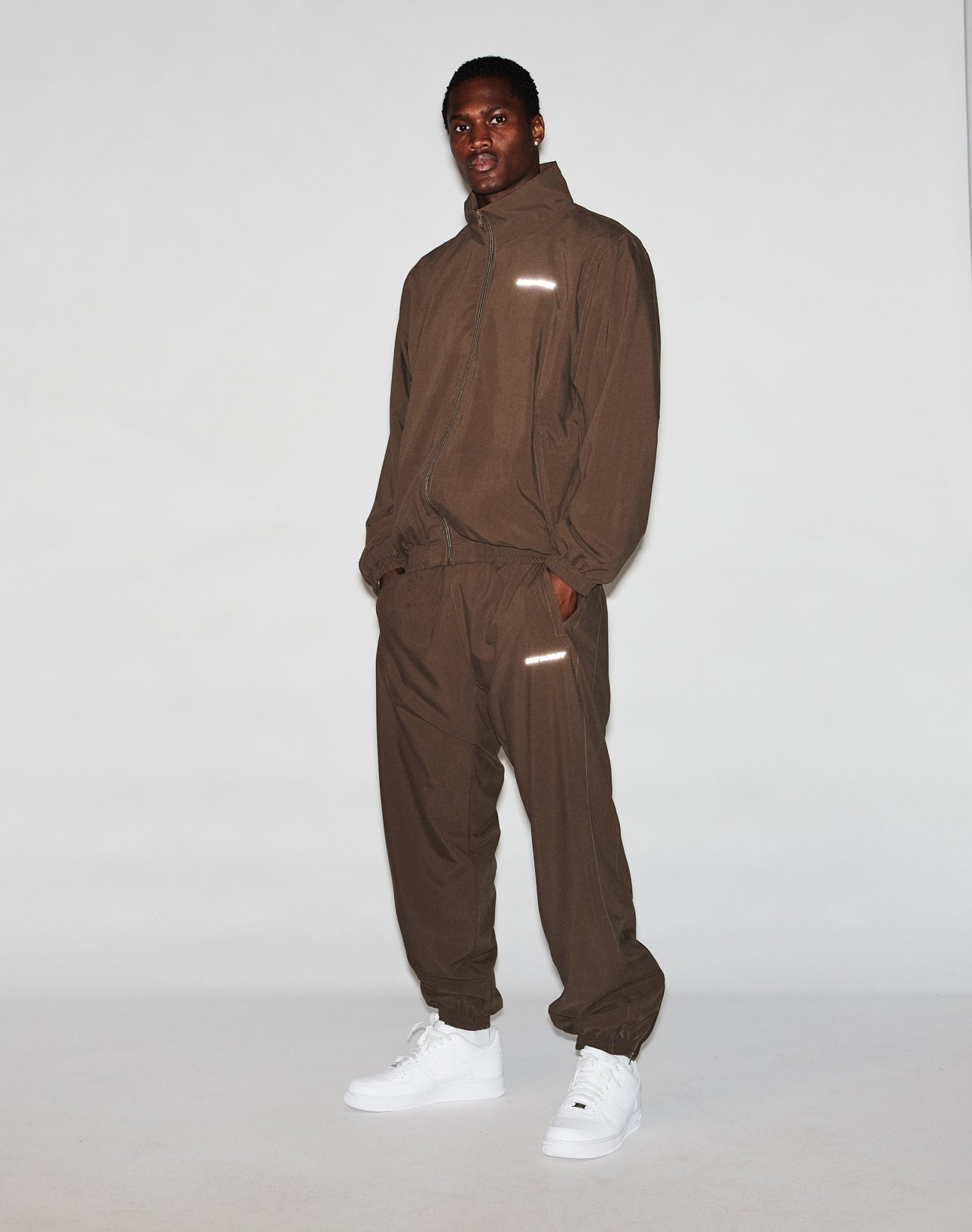 A man poses confidently against a plain background wearing the LIVE FAST DIE YOUNG LFDY Trackjacket and matching pants in an oversized fit, paired with white sneakers. His hands are in his pockets as he looks at the camera.