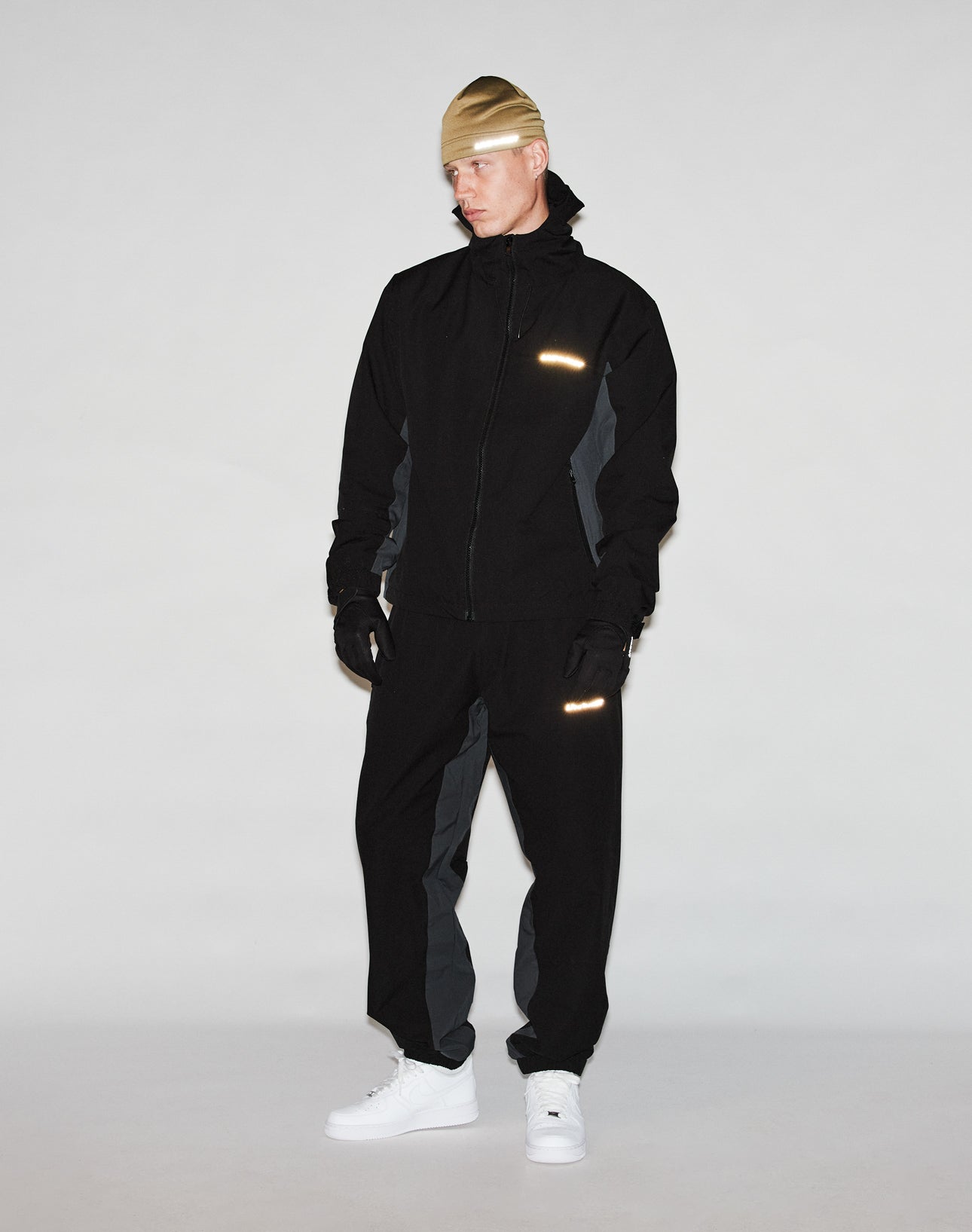 A person wearing a tan beanie, black and gray jacket, pants with reflective print, LIVE FAST DIE YOUNG Live Fast Gloves with touchscreen capability, and white sneakers stands against a plain light background.