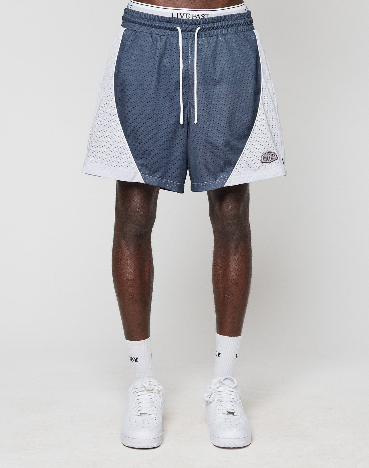 A person stands against a white background wearing LFDY Piping Mesh Shorts by LIVE FAST DIE YOUNG—blue and white polyester with an elastic waistband reading LIVE FAST—plus white crew socks and sneakers. The image is cropped at the waist.