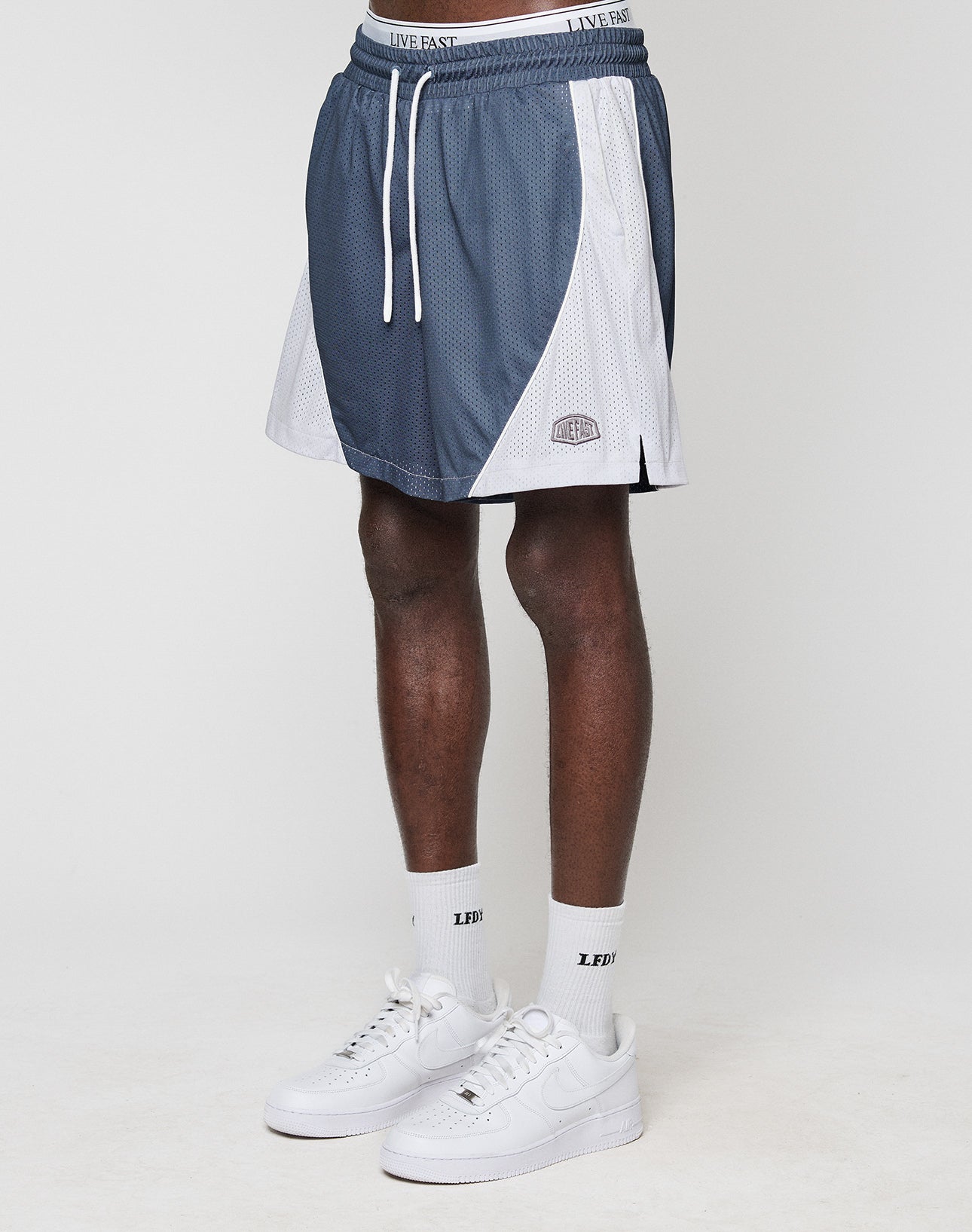 A person wears blue and white LFDY Piping Mesh Shorts by LIVE FAST DIE YOUNG with a LIVE FAST waistband, white socks with LED text, and white Nike sneakers, standing against a plain white background.
