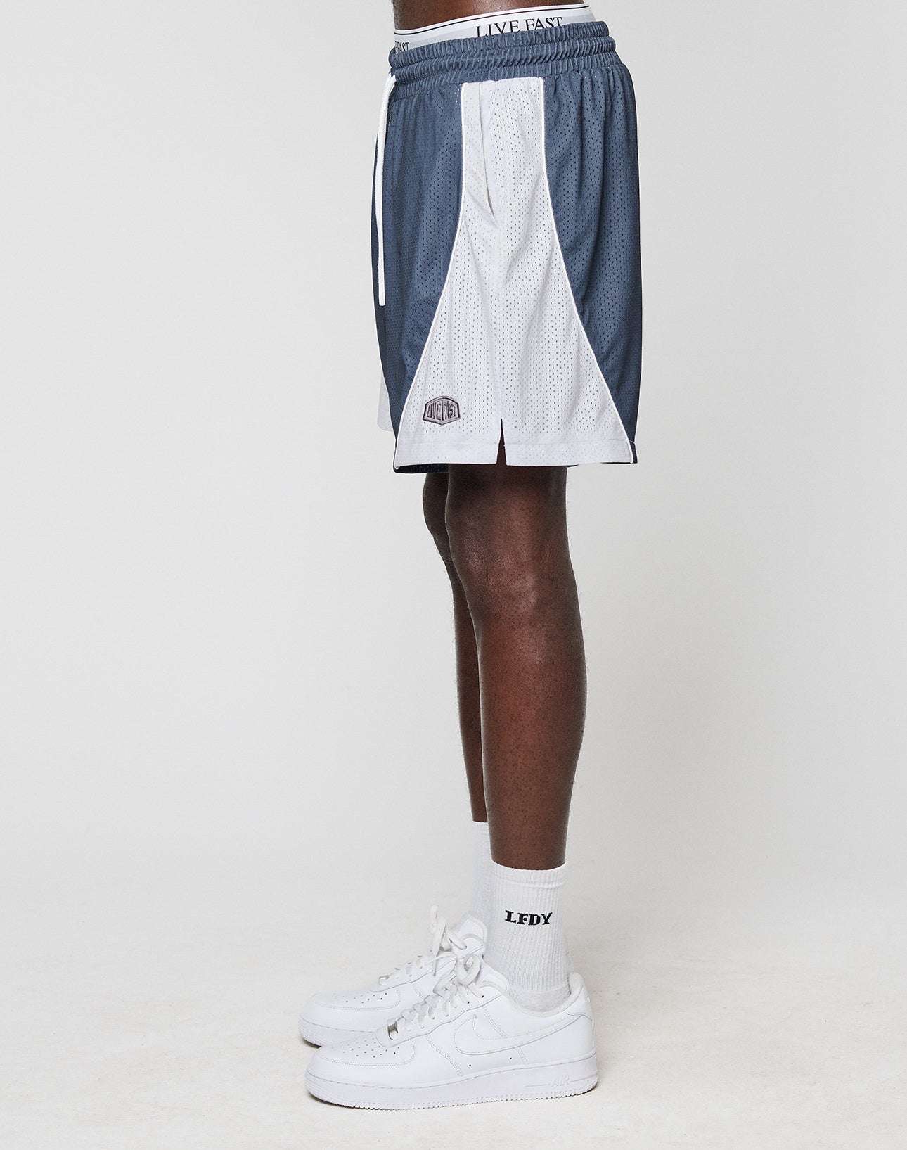 A person wears blue and white LFDY Piping Mesh Shorts by LIVE FAST DIE YOUNG, white crew socks with LIFE and EASY text, and white Nike sneakers, standing sideways against a plain white background.