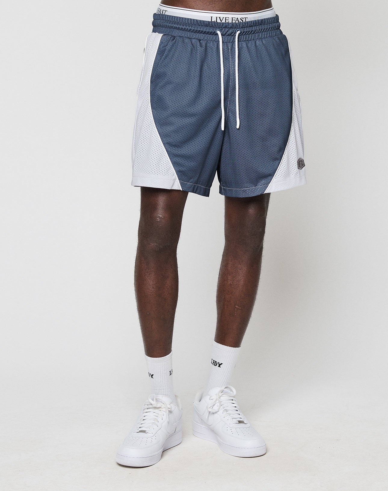 A person stands against a plain white background wearing LIVE FAST DIE YOUNG LFDY Piping Mesh Shorts in blue and white with a regular fit, elastic waistband, drawstring, and mesh side panels. Only the lower body is visible; socks and sneakers are white.