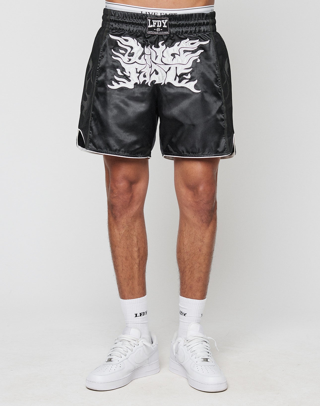 A person wears LFDY Fighting Shorts by LIVE FAST DIE YOUNG—black satin with white flame design, Elastikbund waistband with LFDY logo, smooth polyester, regular fit—paired with white crew socks and sneakers. Plain white background.