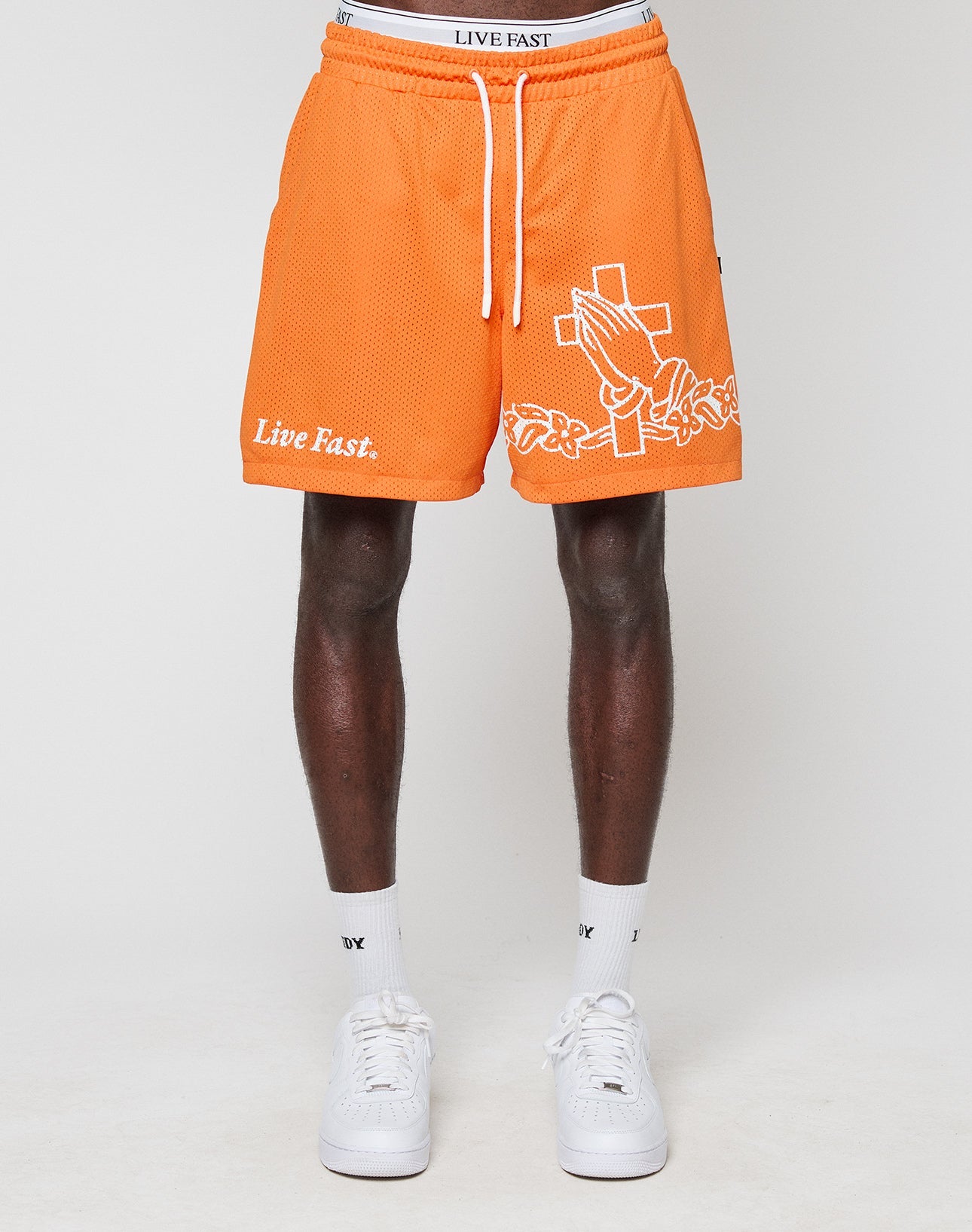 Wearing LIVE FAST DIE YOUNGs LFDY Praying Mesh Shorts in orange, featuring a regular fit, praying hands graphic, cross, and Live Fast text. Made from polyester with an elastic waistband. Paired with white socks and sneakers.