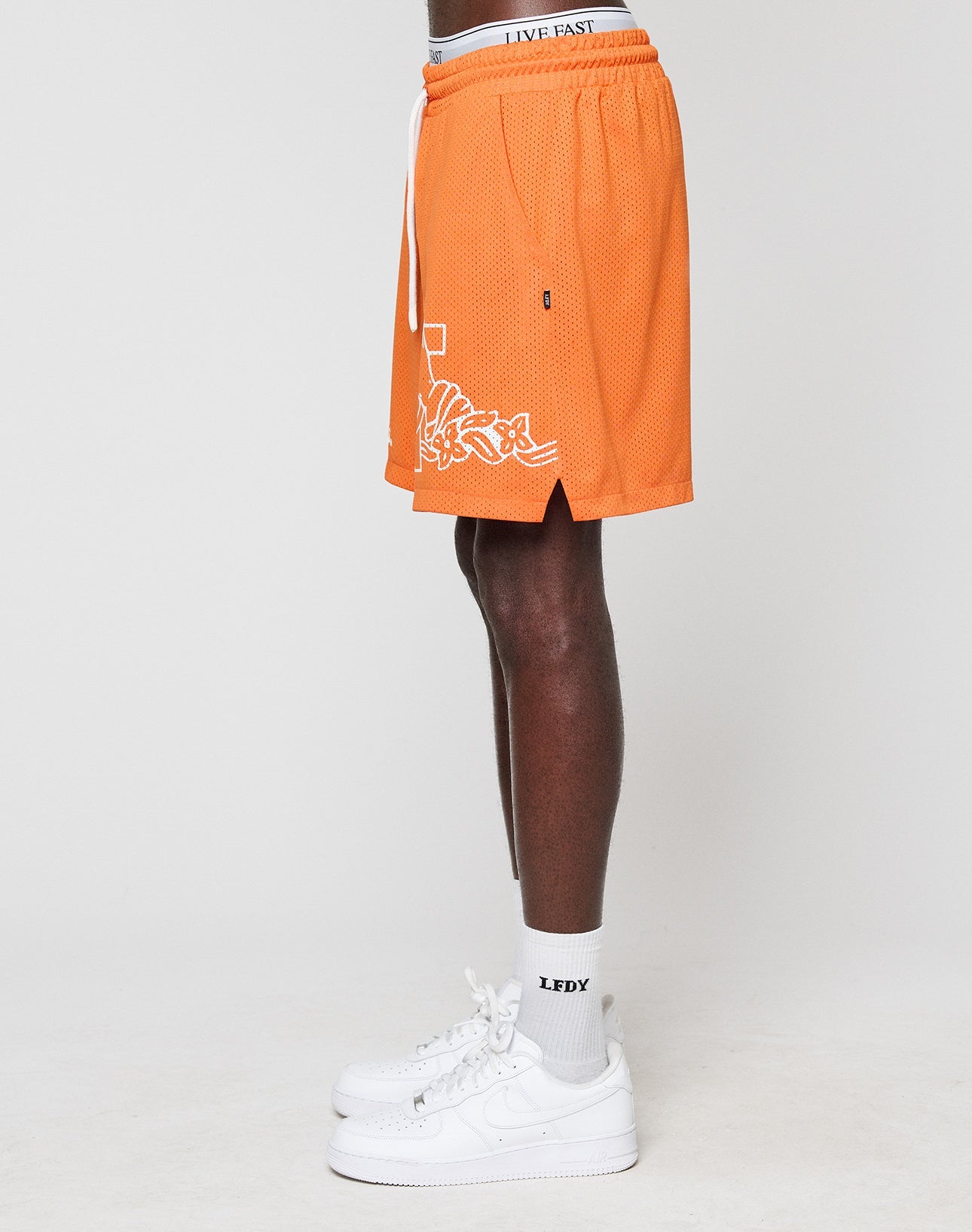 A person stands sideways wearing orange LFDY Praying Mesh Shorts by LIVE FAST DIE YOUNG, featuring white graffiti-style text, a drawstring, and an elastic waistband reading LIVE FAST. They pair them with white LFDY socks and white Nike sneakers.