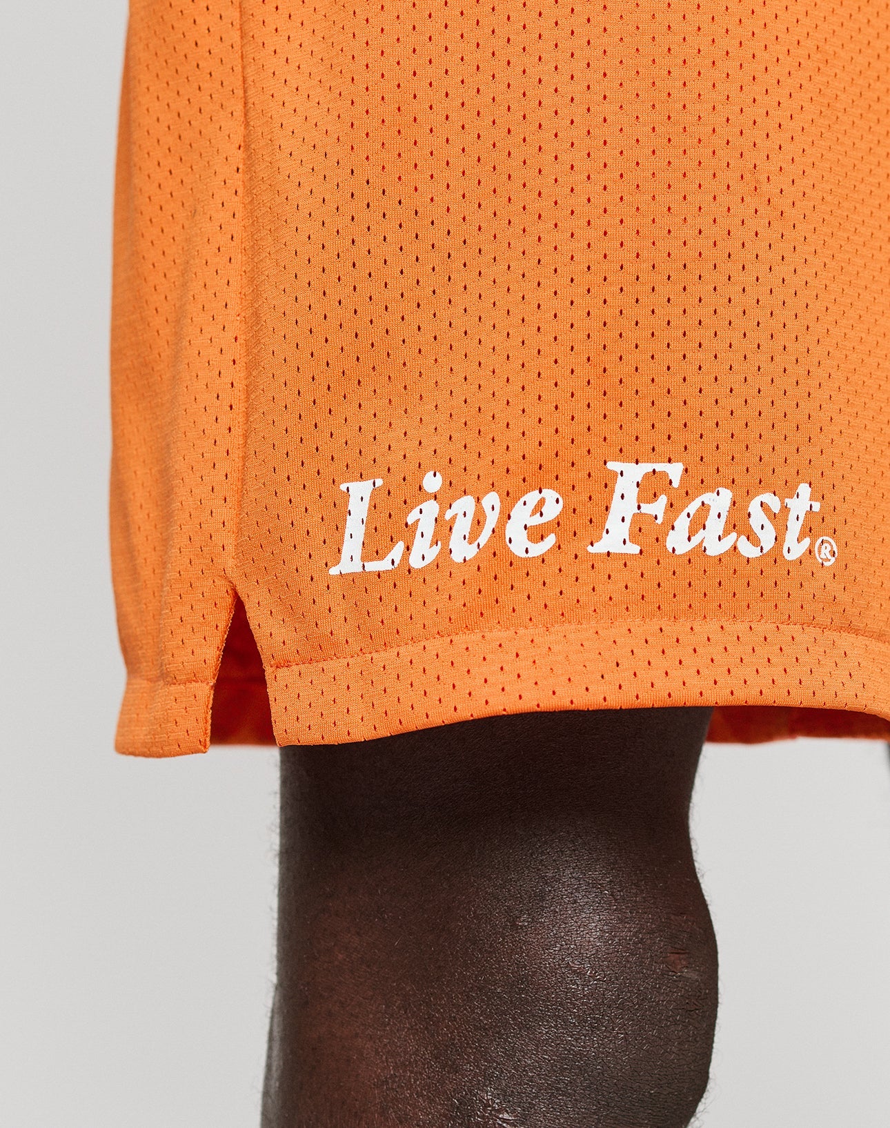 A close-up of a person wearing LFDY Praying Mesh Shorts by LIVE FAST DIE YOUNG, featuring a comfy elastic waistband and Live Fast. in white near the hem. The regular fit orange shorts partially reveal the persons dark-skinned leg.