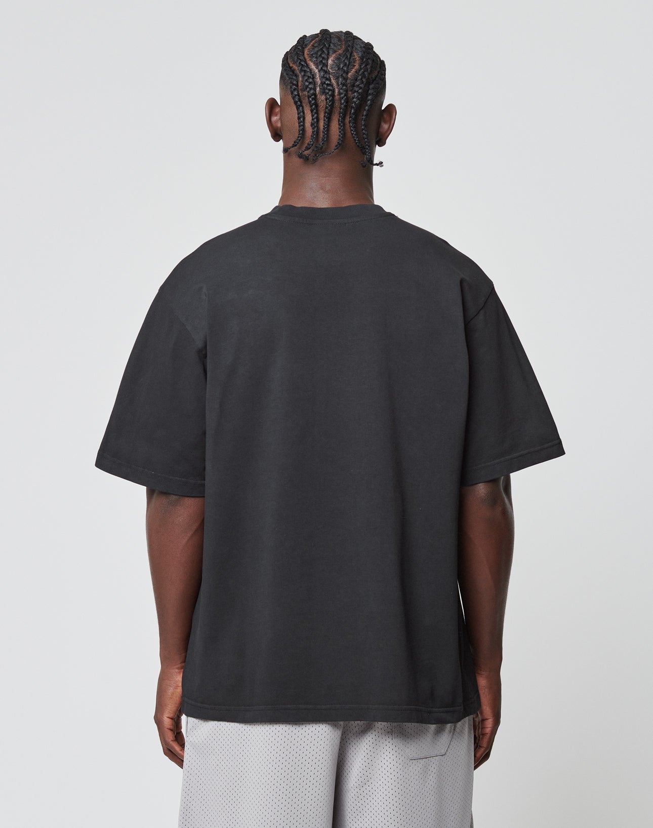A person with braided hair is seen from behind in a LIVE FAST DIE YOUNG Basic Tee, an oversize fit black T-shirt made of 100% Baumwolle, paired with light gray mesh shorts, standing against a plain white background.