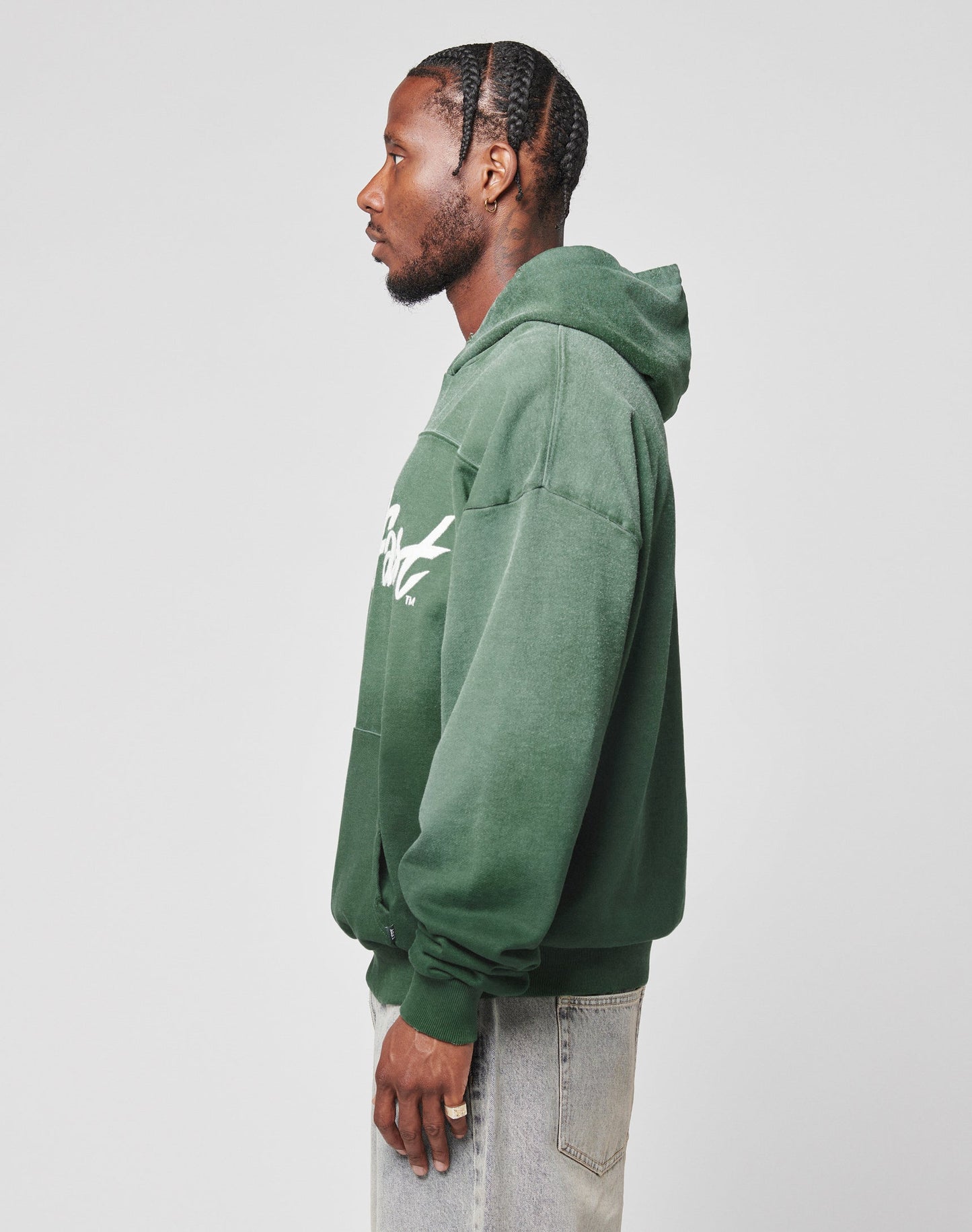 A man with braided hair wears an oversized LFDY Script Hooded by LIVE FAST DIE YOUNG in green and light-wash jeans, standing in profile against a plain white background.