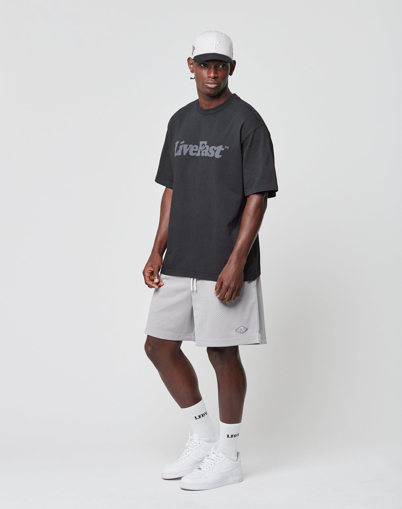A man stands against a plain background wearing a black Basic Tee by LIVE FAST DIE YOUNG (Vintage-Waschung, Oversize Fit), light gray cap and shorts, white LIVE socks, and white sneakers. He faces forward with a neutral expression.