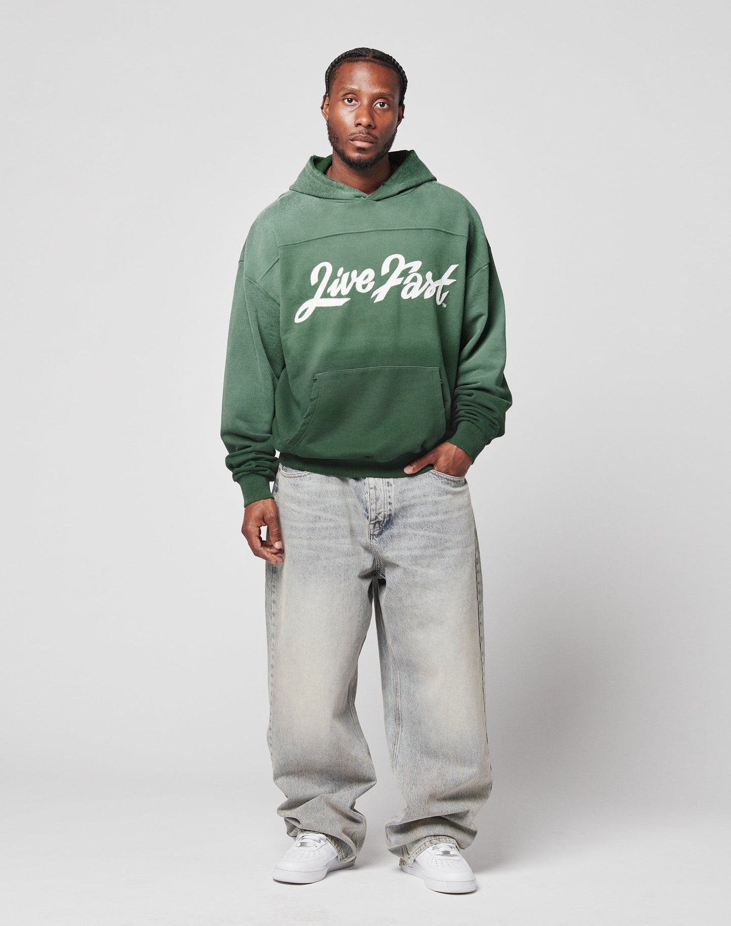 A person stands against a plain background, wearing the LIVE FAST DIE YOUNG LFDY Script Hooded in green with Live Fast Sprayeffekt, light gray distressed baggy jeans, and white sneakers. One hand is in their pocket as they face the camera.