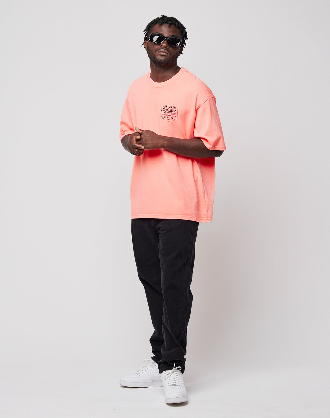 A man with short, twisted hair and sunglasses wears a loose pink LIVE FAST DIE YOUNG Regular Denim T-shirt, black pants, and white sneakers. He stands against a plain white background, relaxed, with one hand touching his shirt.