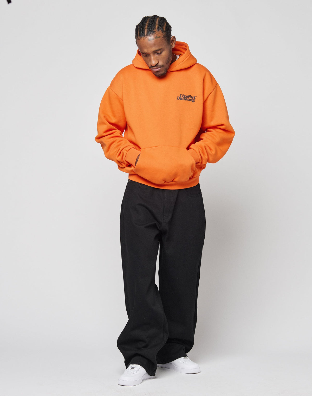 A person stands against a plain background, wearing the LIVE FAST DIE YOUNG Basic Orange Hooded with black front and back print, loose black pants, and white sneakers. They look down with hands in the hoodies pocket.