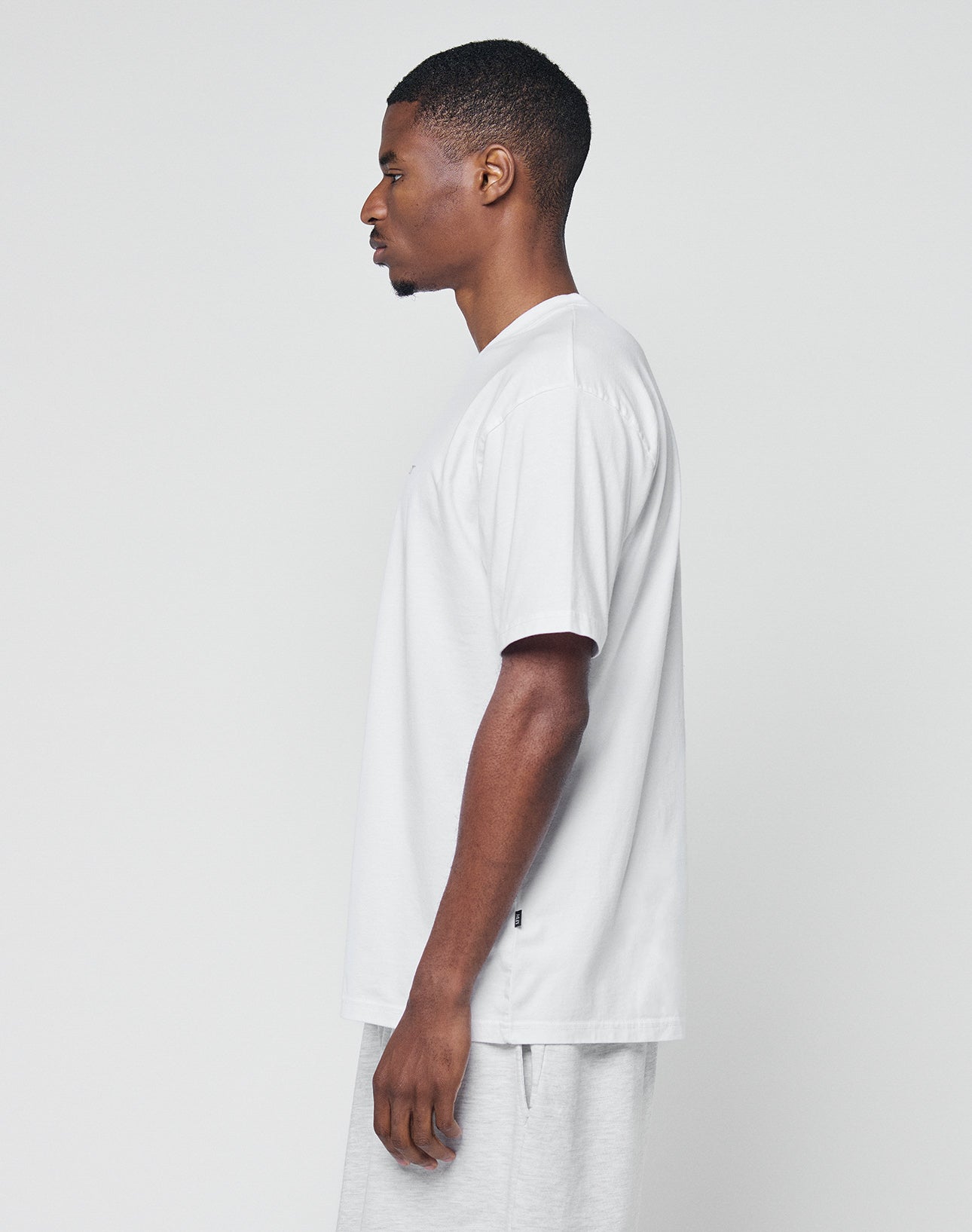 A man stands in profile facing left, wearing the LIVE FAST DIE YOUNG Basic Chest Tee—regular fit, plain white, 100% Baumwolle—and light gray shorts against a plain light background.