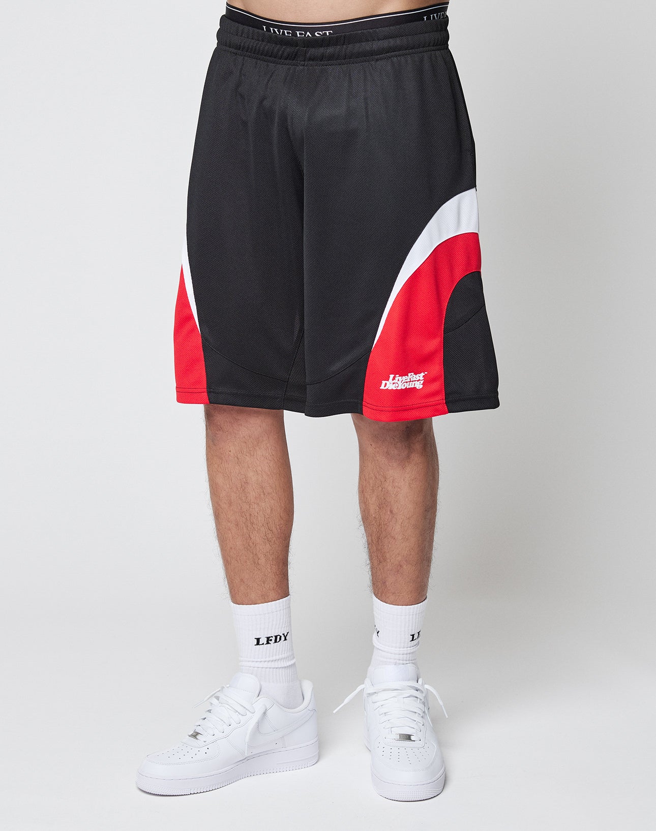 A person in LIVE FAST DIE YOUNG Basic Trackshorts—black polyester with white and red curved accents—paired with white LEDY socks and white sneakers, stands against a plain light background. The modern shorts have an oversize fit; upper body not visible.
