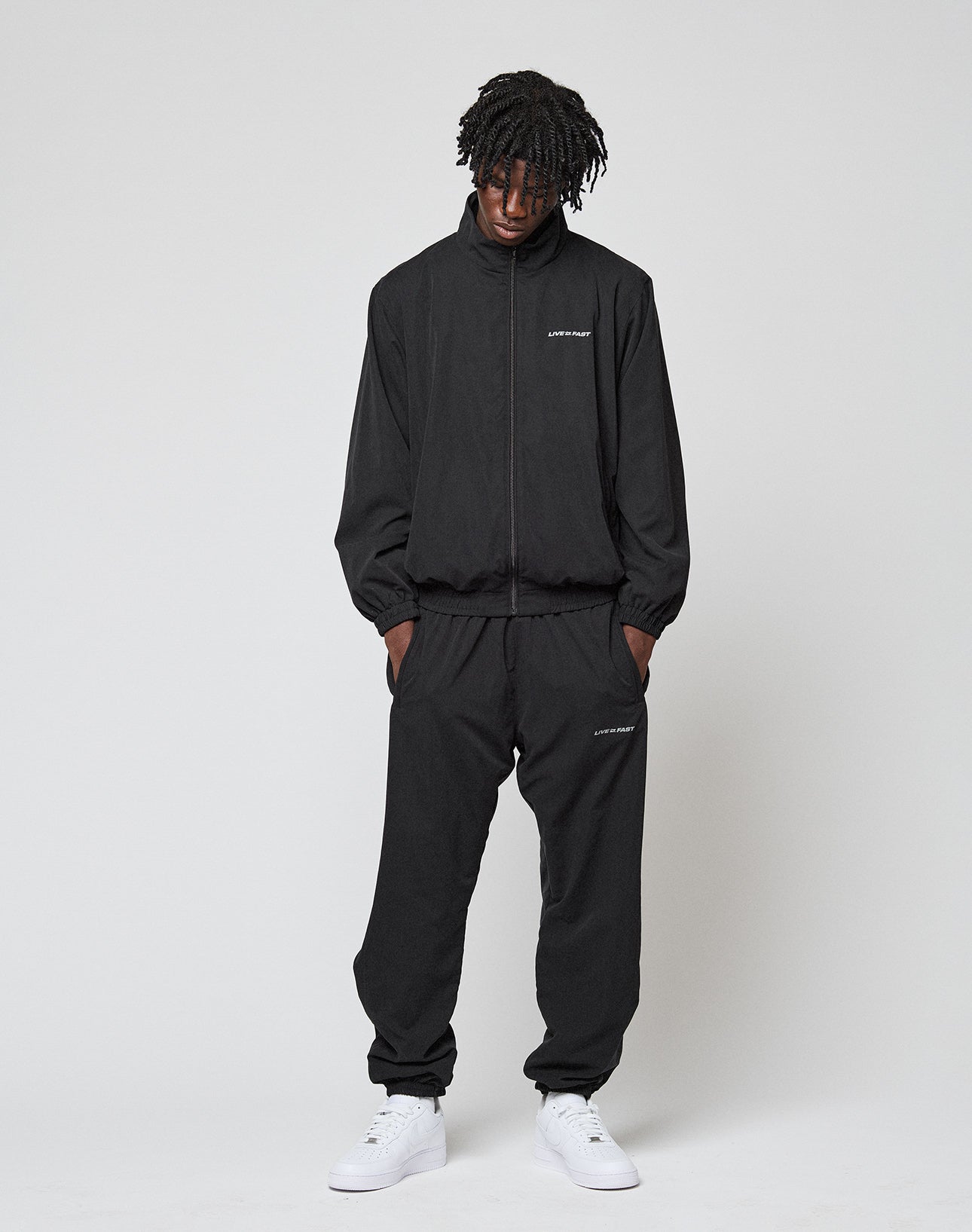 A person stands against a white background wearing the LIVE FAST DIE YOUNG LFDY Trackjacket, an oversized black zip-up with subtle logo details on chest and thigh, paired with white sneakers. Hands in pockets, they look down.