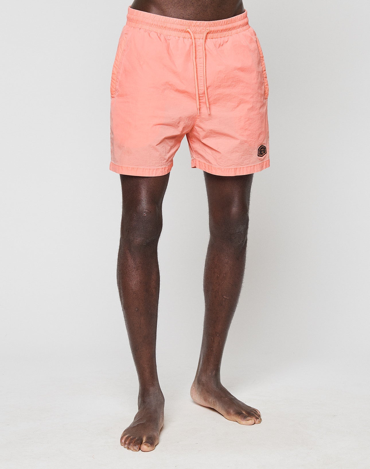 A person with dark skin stands barefoot on a white background, wearing LIVE FAST DIE YOUNGs Live Fast Swim Shorts in light coral pink with an elastic waistband, drawstring, and a small circular patch near the left hem. Upper body not visible.