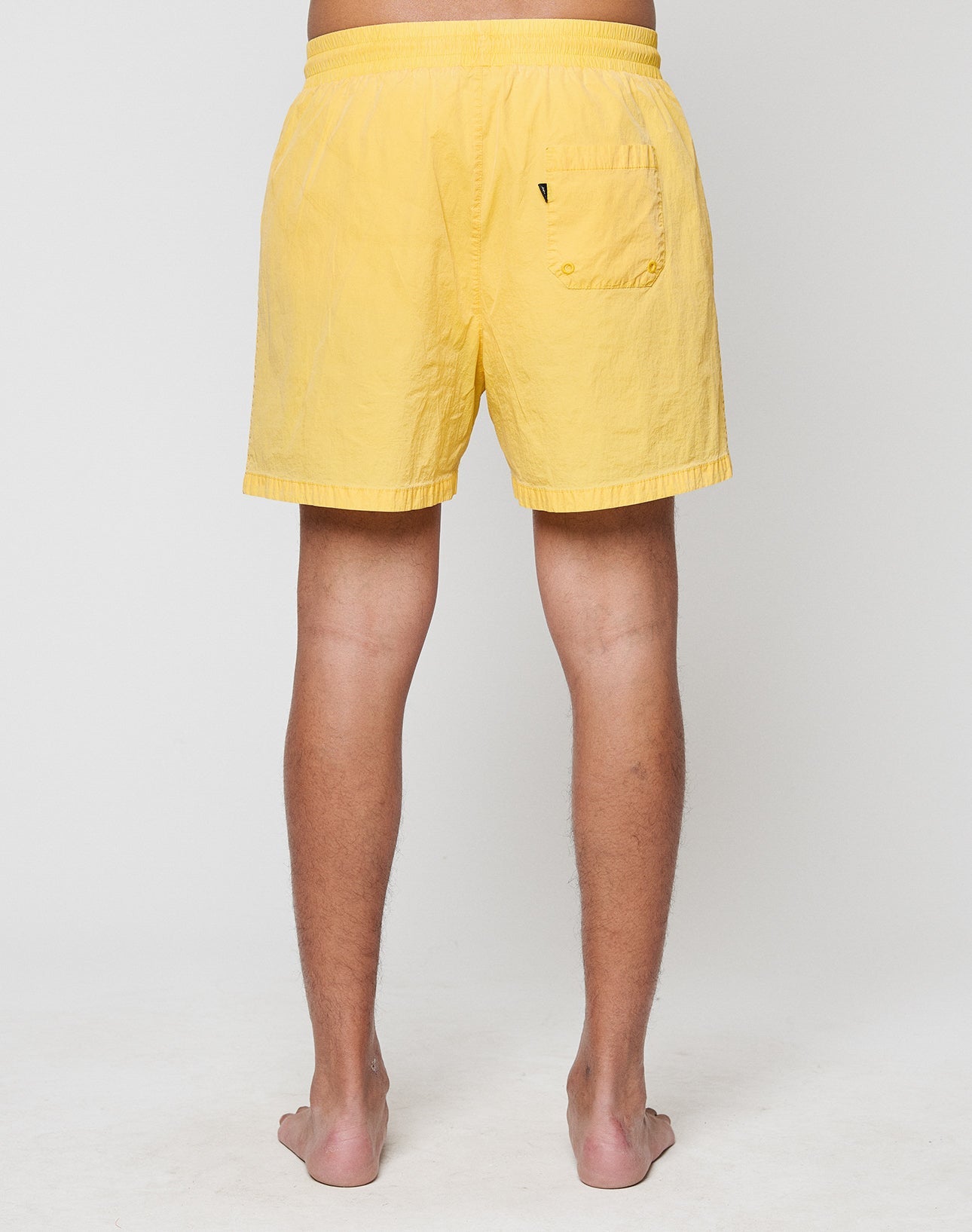 A person stands barefoot facing away from the camera, wearing LIVE FAST DIE YOUNG Live Fast Swim Shorts in a tinted yellow wash with a breiter Elastikbund and single back pocket, set against a plain, light background.