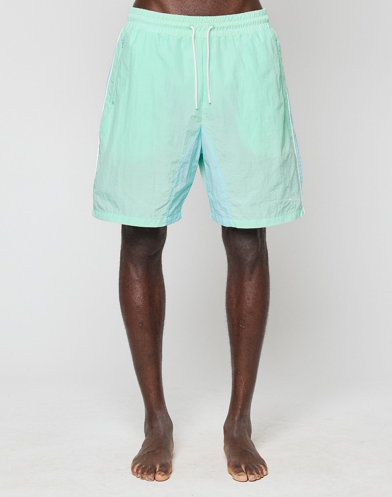 A person stands barefoot on a white surface, wearing mint green LIVE FAST DIE YOUNG Beach Shorts. The image is cropped from the waist to the feet.