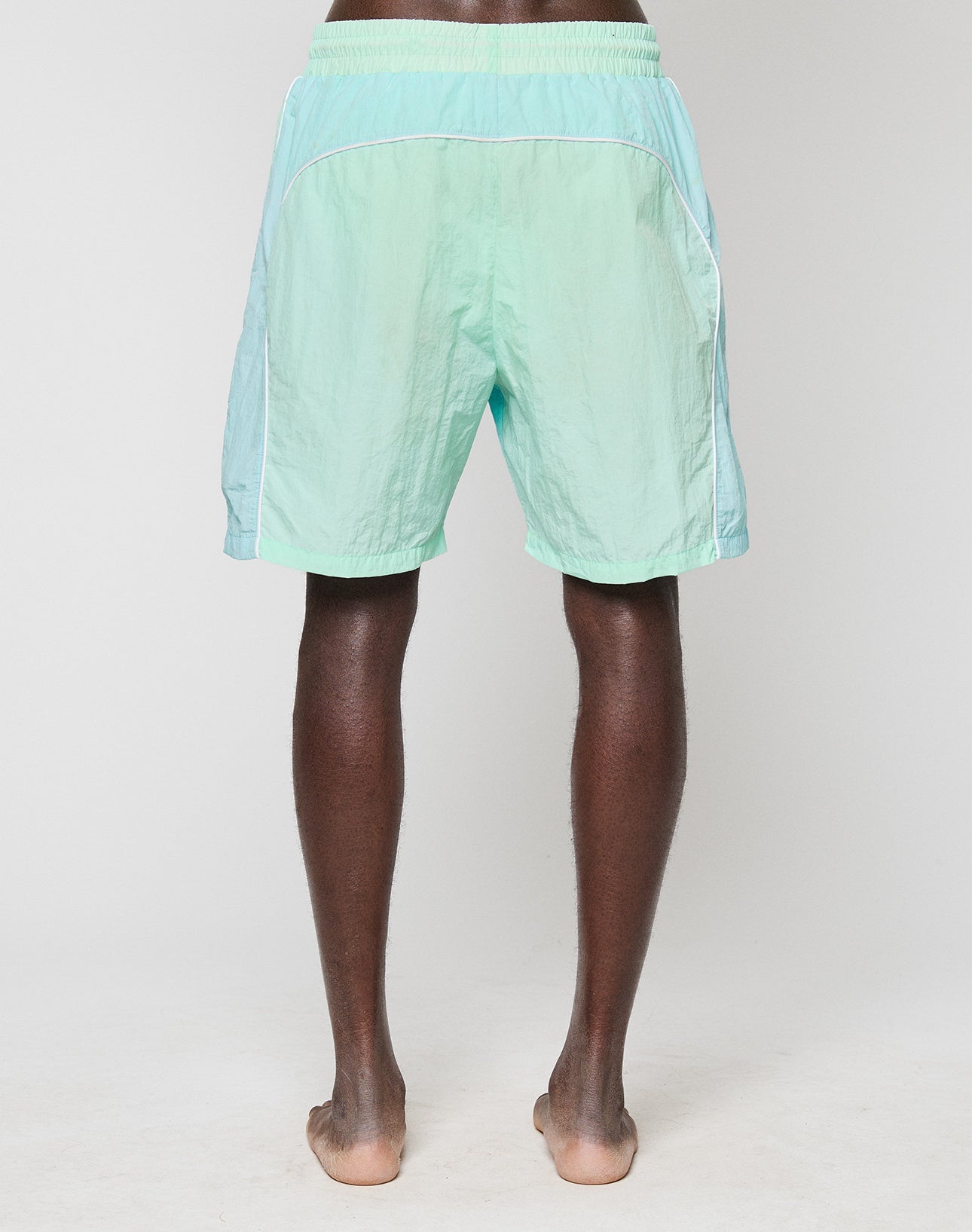 A person stands barefoot on a white surface, shown from the back, wearing LIVE FAST DIE YOUNG Beach Shorts in a light teal and mint green colorway with white piping detail. The background is plain and white.