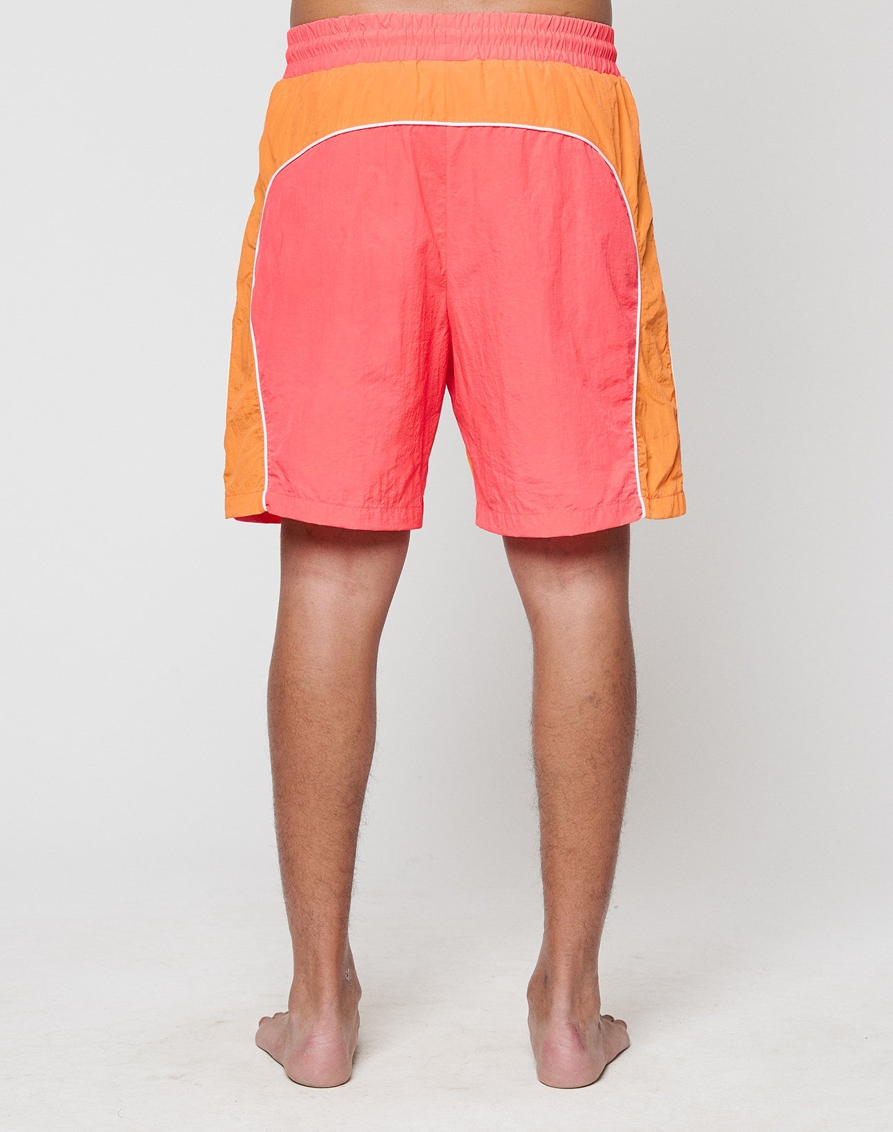 A person stands barefoot, facing away, wearing LIVE FAST DIE YOUNG Beach Shorts in bright orange and coral with white piping and a wide elastic waistband, set against a plain light background.