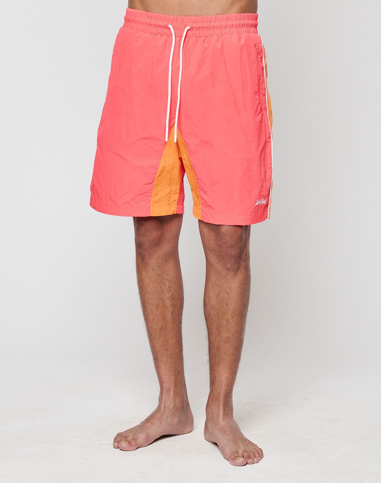A person stands barefoot in LIVE FAST DIE YOUNG Beach Shorts, coral pink with yellow side panels and a wide elastic waistband, against a plain light background. Only the lower torso and legs are visible.