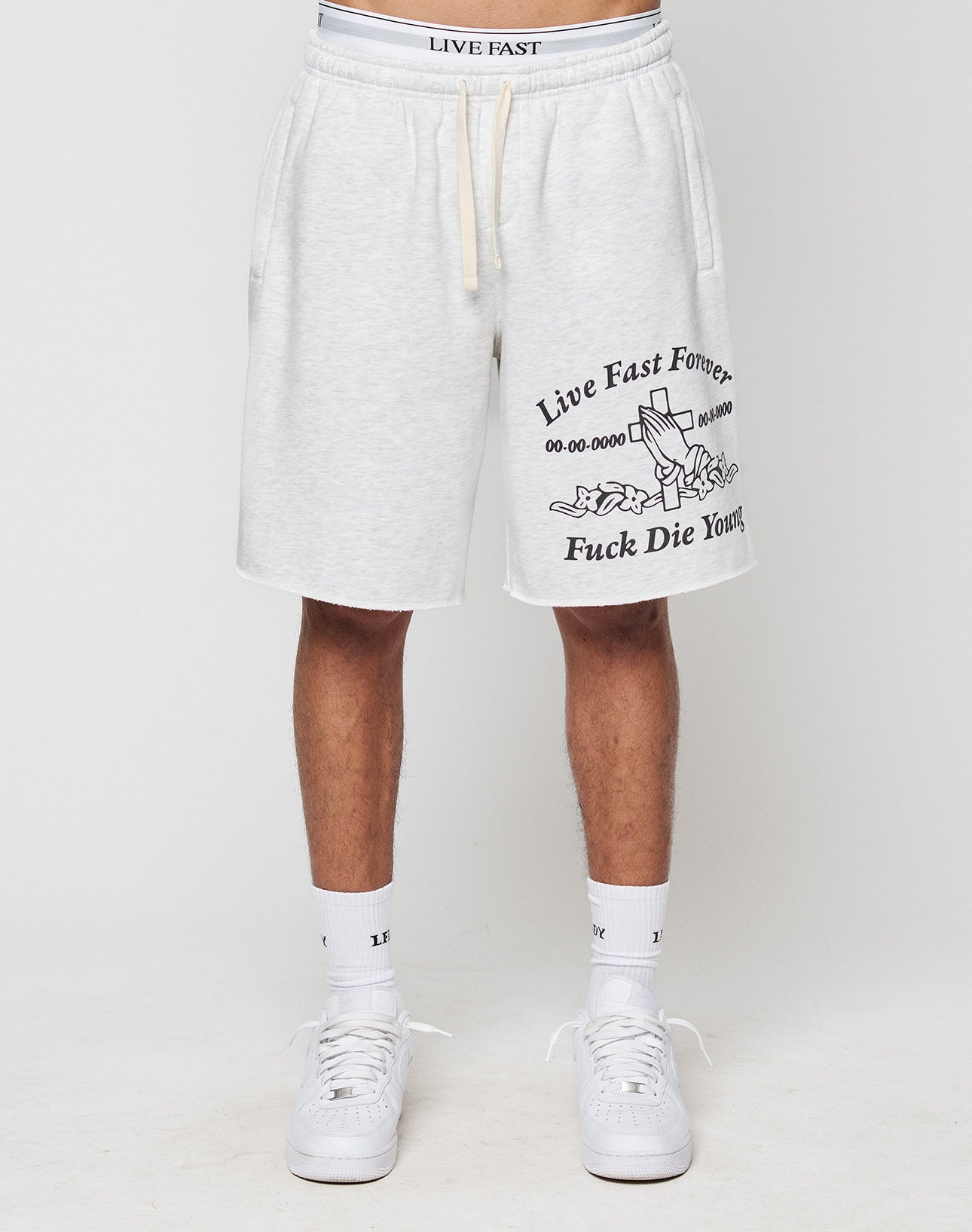 A person wears light gray LFDY Praying Shorts by LIVE FAST DIE YOUNG, featuring black “Live Fast Forever” and “Fuck Die Young” prints, a tombstone graphic, Elastikbund, seitliche Eingrifftaschen, white socks, and sneakers.