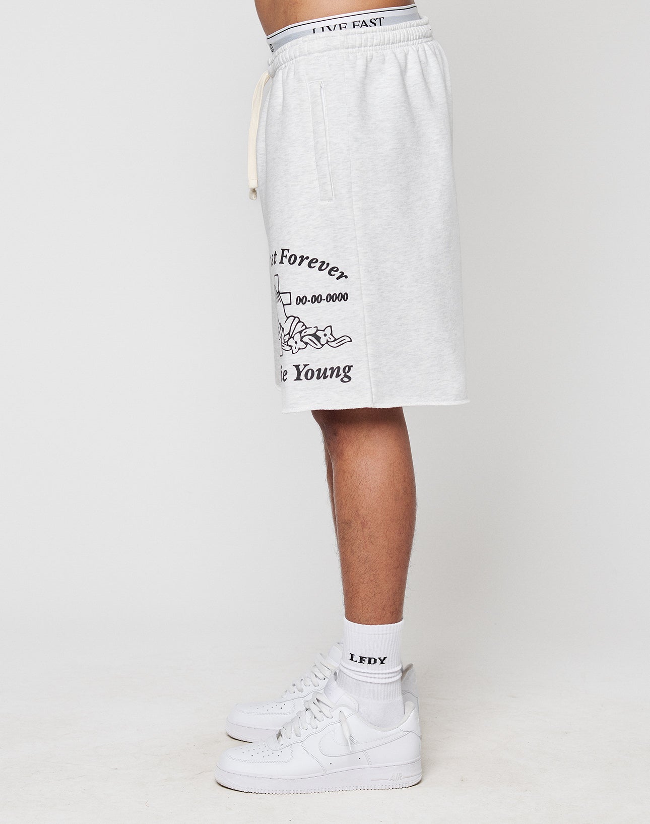 A person stands sideways in LFDY Praying Shorts by LIVE FAST DIE YOUNG, light gray with black graphic text and illustration on the left leg, featuring seitliche Eingrifftaschen, Elastikbund, and paired with white crew socks and sneakers. Plain white background.