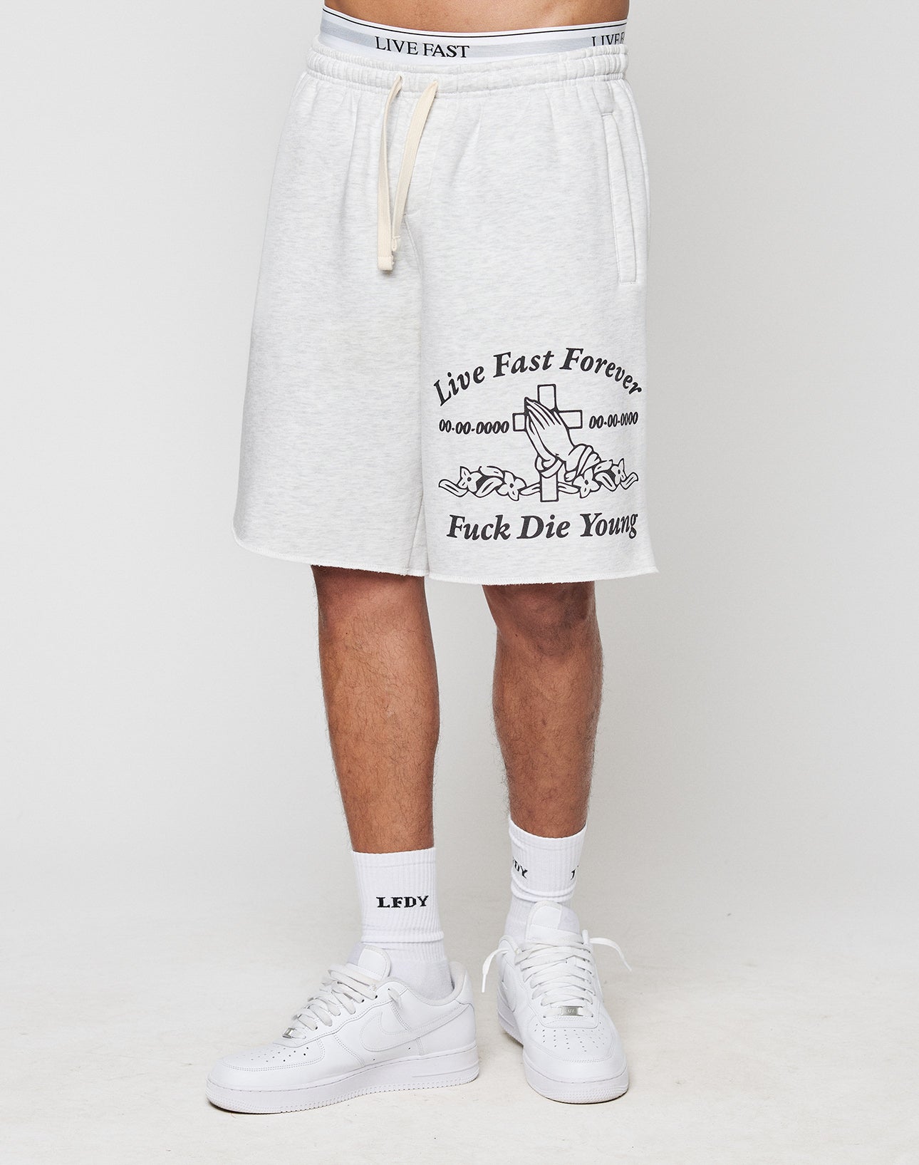 Someone wears light gray LFDY Praying Shorts by LIVE FAST DIE YOUNG with elastic waistband, side pockets, urn and flower graphic, Live Fast Forever Fuck Die Young text, white LFDY socks, white sneakers, and a matching LIVE FAST waistband.