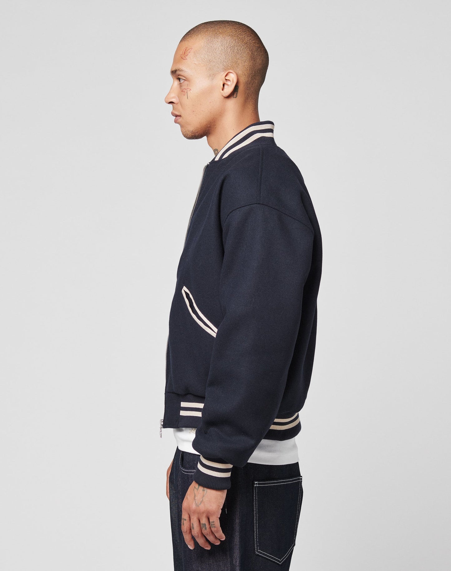 A person with a shaved head stands in profile against a plain background, wearing the Live Fast College Jacket by LIVE FAST DIE YOUNG, featuring a DY-Reißverschluss Puller and Rückenstick Gefüttert, paired with regular-fit dark jeans.
