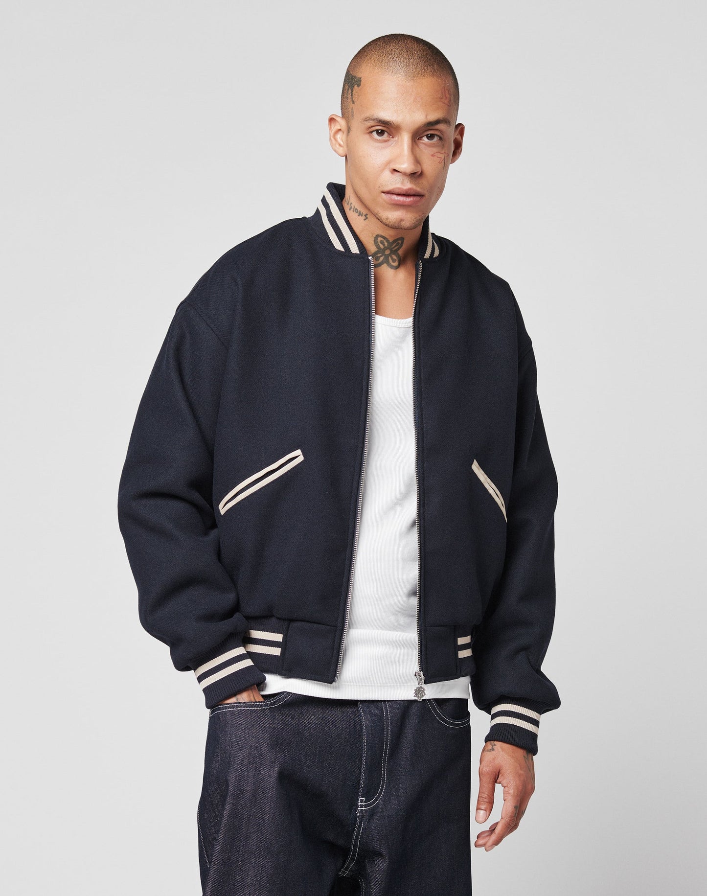 A person with a shaved head and neck tattoo wears the LIVE FAST DIE YOUNG Live Fast College Jacket with cream stripes over a white shirt and dark jeans, standing against a plain light background.