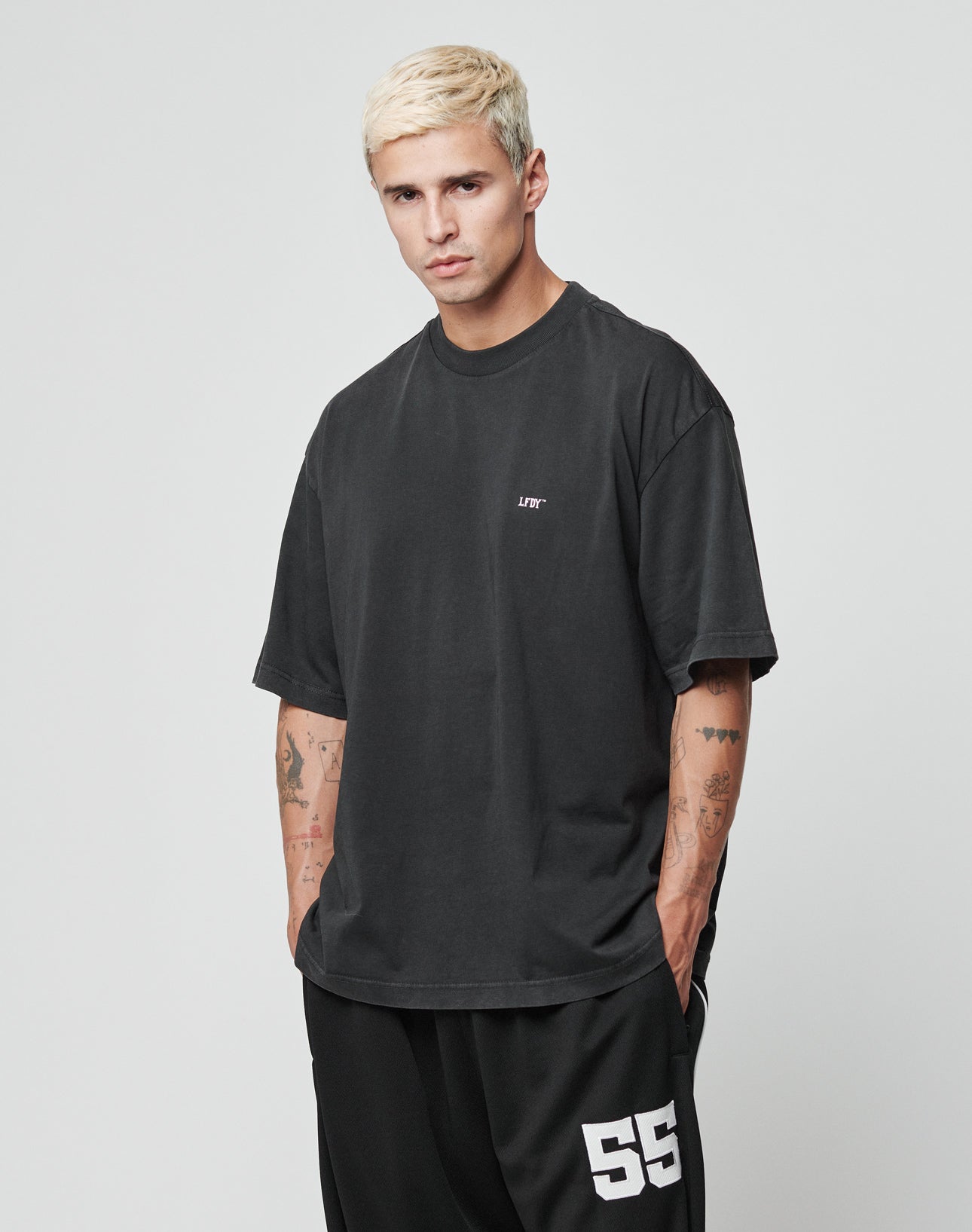 A young man with short bleached blond hair wears the LIVE FAST DIE YOUNG LFDY Flip Phone Tee in an oversized black cotton fit with small white text and black shorts numbered 55, standing with hands in pockets against a plain light background.