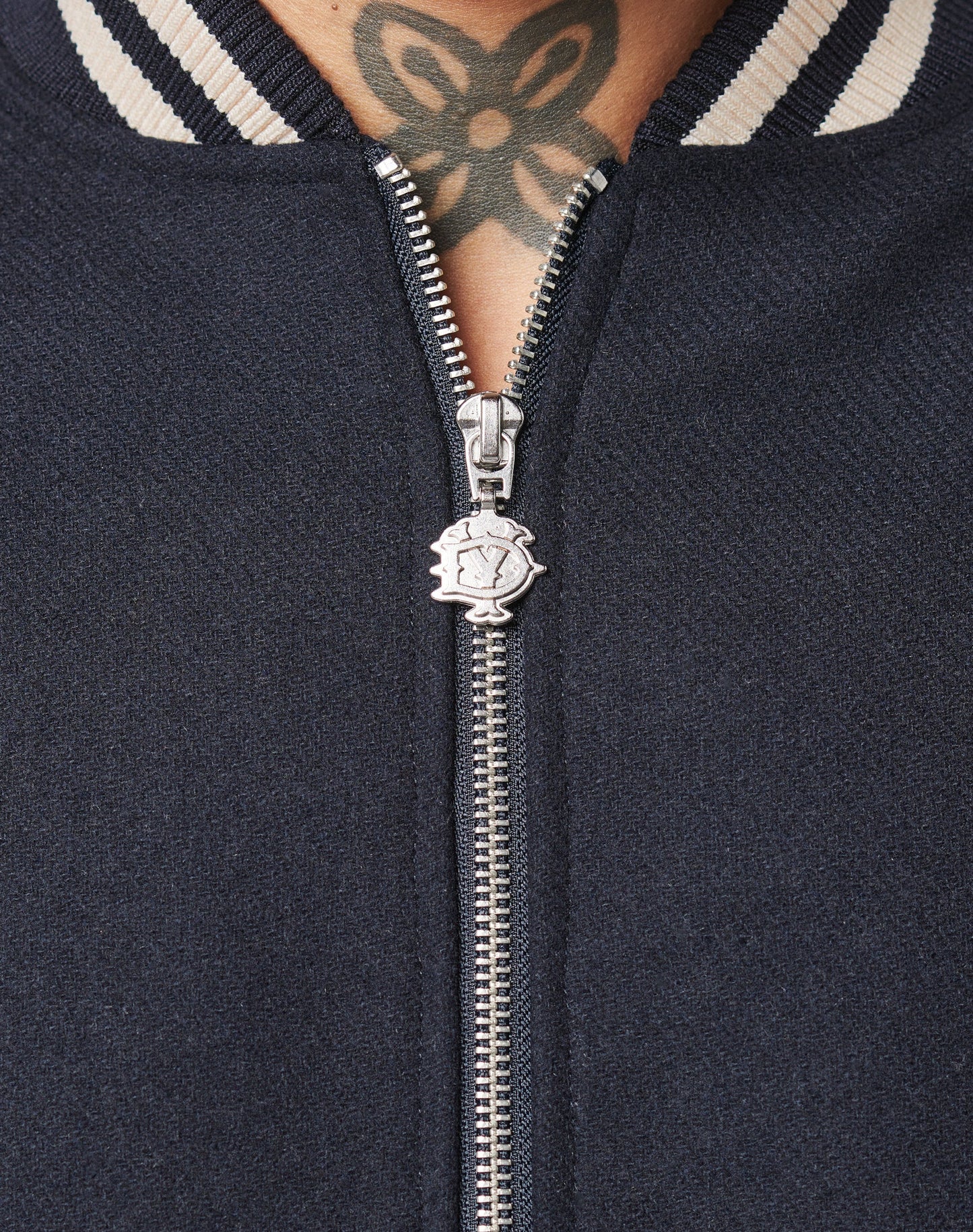 Close-up of a silver DY-Reißverschluss Puller on the navy Live Fast College Jacket by LIVE FAST DIE YOUNG, featuring a regular fit and white/navy striped collar. A black floral tattoo appears on the wearer’s upper chest and neck above the collar.