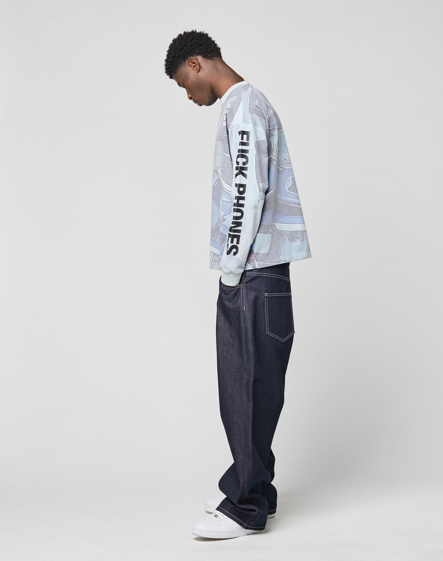 A young man in profile wears the LIVE FAST DIE YOUNG LFDY Stolen Phones Longsleeve, oversize fit in light blue with FUCK PHONES sleeve print, wide vintage-wash dark jeans, and white sneakers against a plain light background.