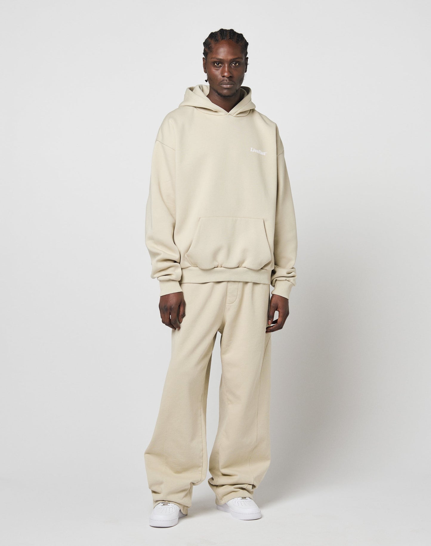 A person poses against a plain white background, wearing the LIVE FAST DIE YOUNG Basic 360 Hooded beige oversized hoodie and matching loose-fit sweatpants made of soft Baumwolle, paired with white sneakers, looking directly at the camera.