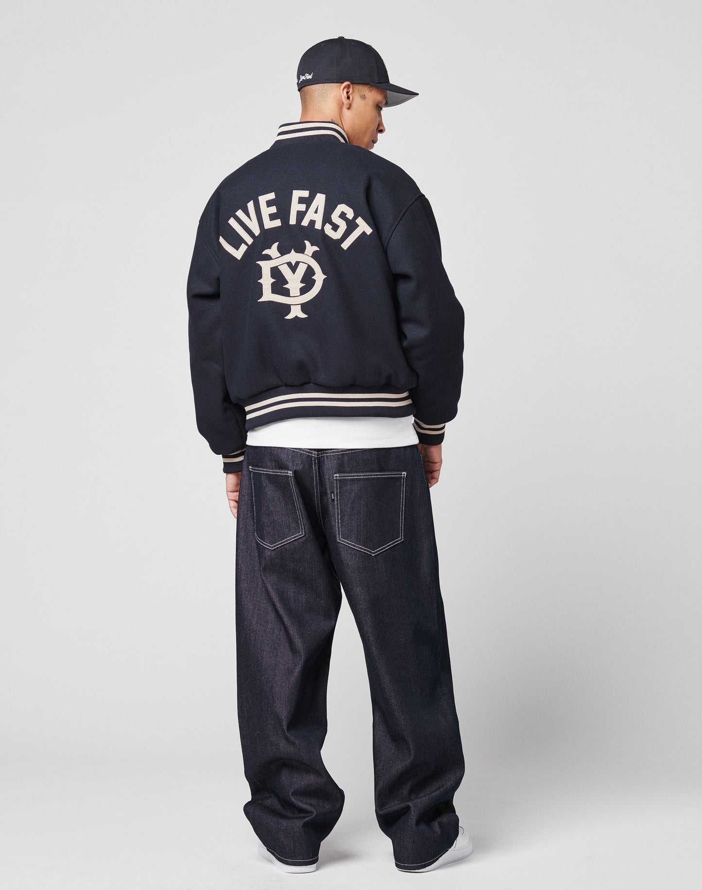 A person stands facing away, wearing the LIVE FAST DIE YOUNG Live Fast College Jacket with Rückenstick logo, a black cap, and loose-fitting dark jeans against a plain, light background.