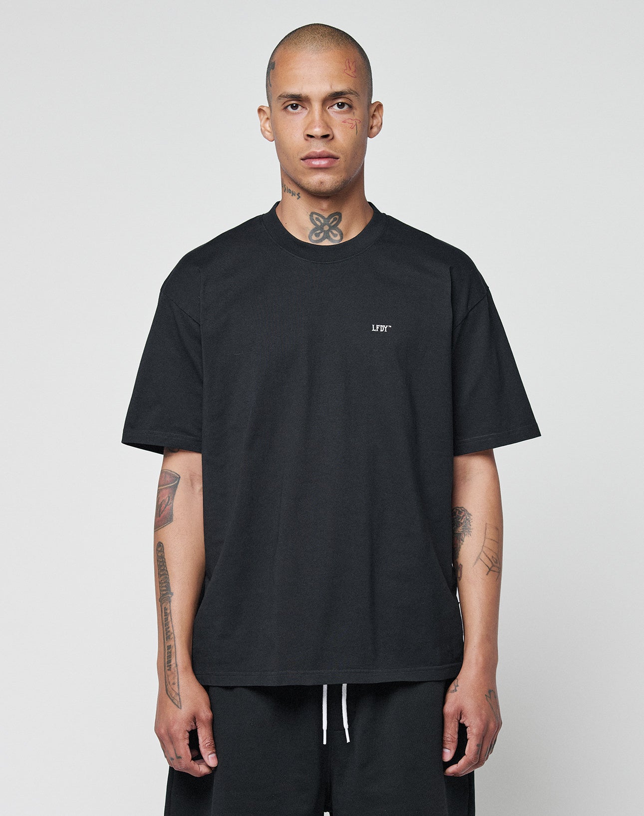 A person with short hair and visible neck and arm tattoos stands facing forward in a neutral pose, wearing the LIVE FAST DIE YOUNG Basic Chest Tee (100% Baumwolle) in black, paired with black shorts against a plain background.