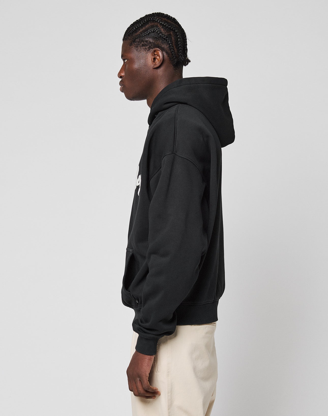 A person with braided hair stands in profile facing left, wearing the LIVE FAST DIE YOUNG Monaco Boys Hooded black hoodie in an oversize fit crafted from a Baumwolle-Polyester mix, paired with light pants against a plain light gray background.