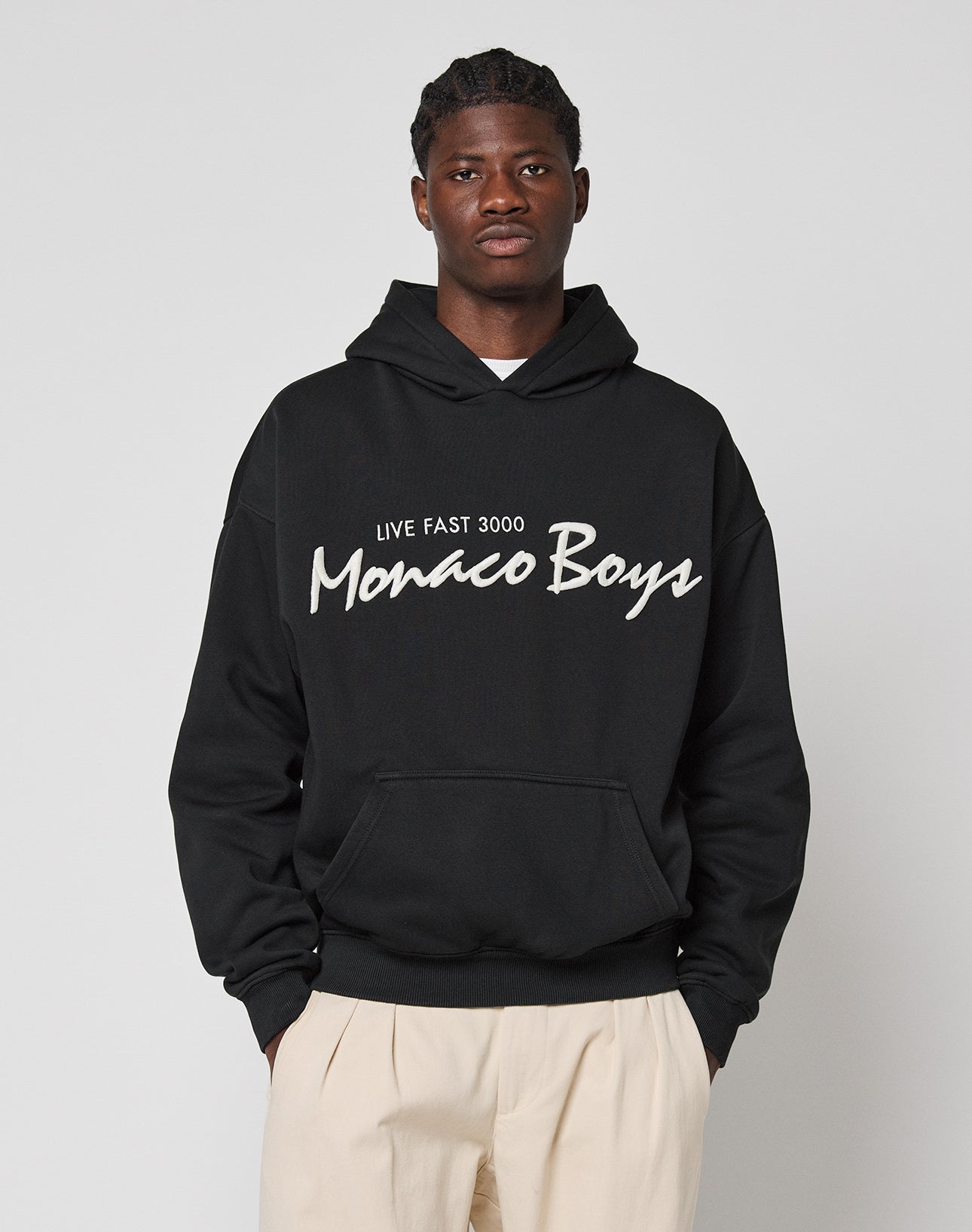 A man in an oversized black Monaco Boys Hooded by LIVE FAST DIE YOUNG stands against a light gray background. He has short hair and wears light-colored pants made of a soft cotton-polyester blend.