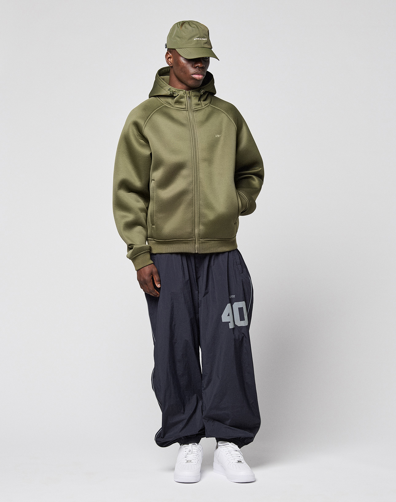 A man stands against a plain background wearing the LFDY Scuba Hooded Zip by LIVE FAST DIE YOUNG, loose dark gray pants with a white "40" on the leg, and white sneakers for a comfortable Regular Fit look.