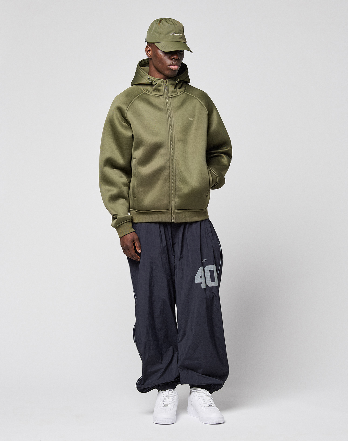 A man stands against a plain background wearing the LFDY Scuba Hooded Zip by LIVE FAST DIE YOUNG, loose dark gray pants with a white "40" on the leg, and white sneakers for a comfortable Regular Fit look.