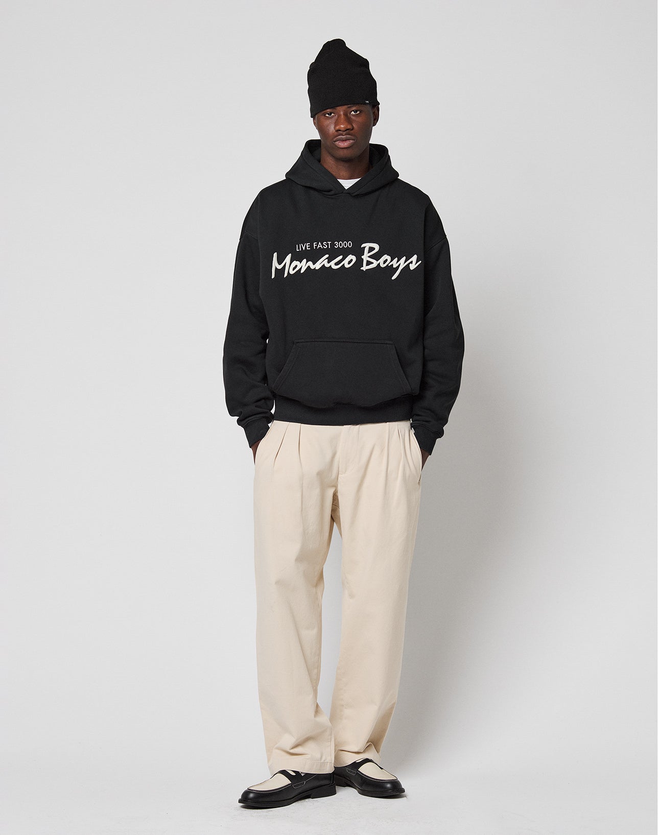 A person stands against a plain background wearing a black beanie, beige pleated trousers, black-and-white dress shoes, and an oversize fit LIVE FAST DIE YOUNG Monaco Boys Hooded in a cotton-polyester mix, hands in pockets and neutral expression.