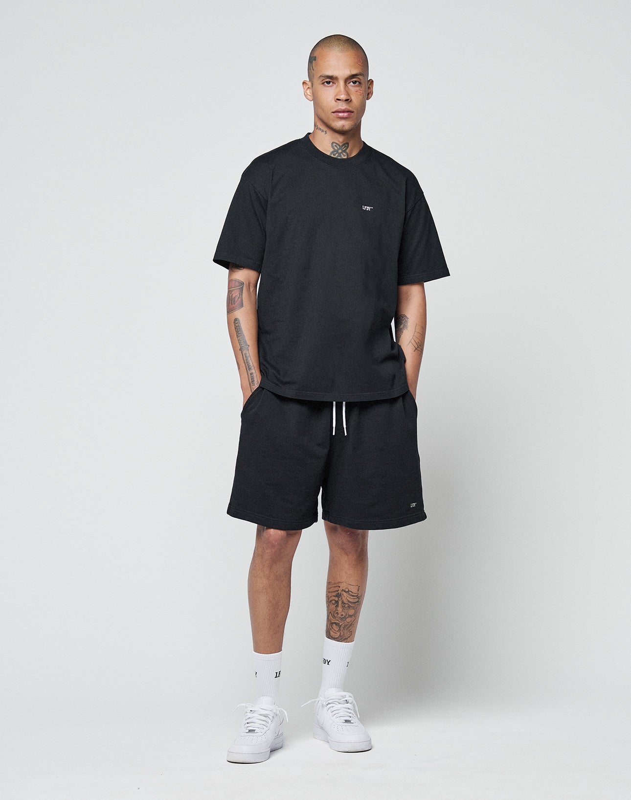 A person stands against a white background wearing the LIVE FAST DIE YOUNG Basic Chest Tee in black, paired with black shorts, white crew socks, and white sneakers. Their hands are in their pockets, showing tattoos on their arms and legs.